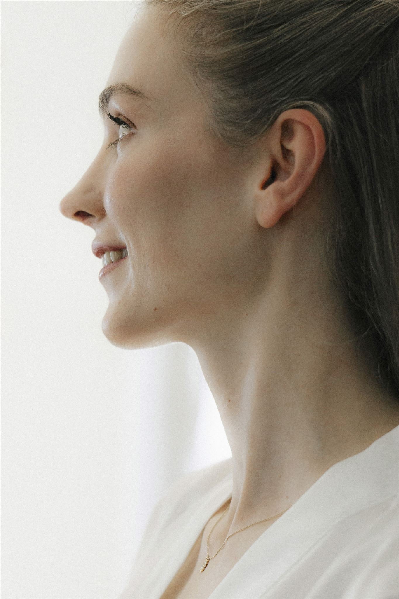 Nahaufnahme einer lächelnden Braut von der Seite, mit braunen Haaren, Schmuck und heller Haut, vor einem weißen Hintergrund.