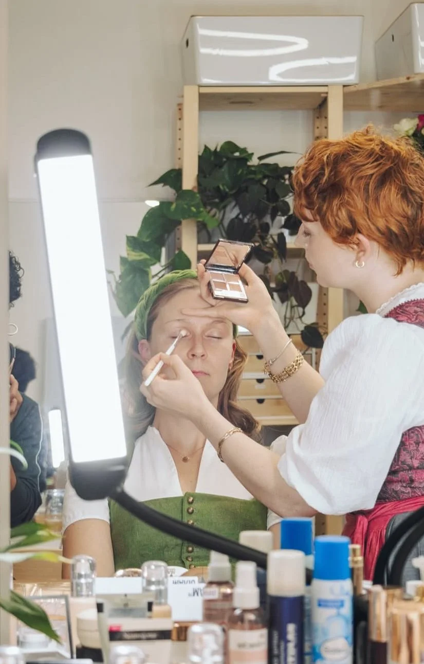 Eine Frau wird von einer Make-up Artistin geschminkt, im Hintergrund Pflanzen und Make-up-Produkte auf dem Tisch.