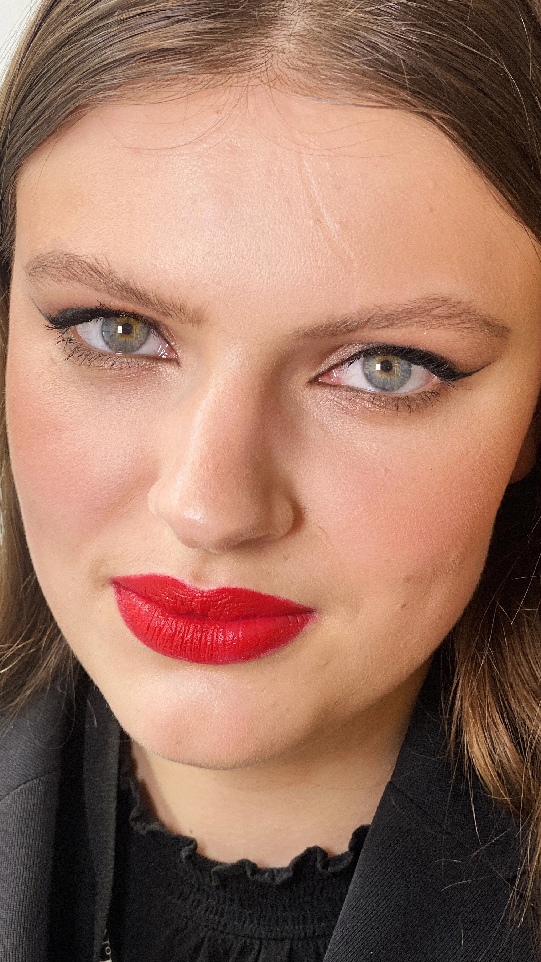 Nahaufnahme eines Gesichts einer jungen Frau mit rotem Lippenstift, heiteren Augen, dunklem Eyeliner und braunem Haar.