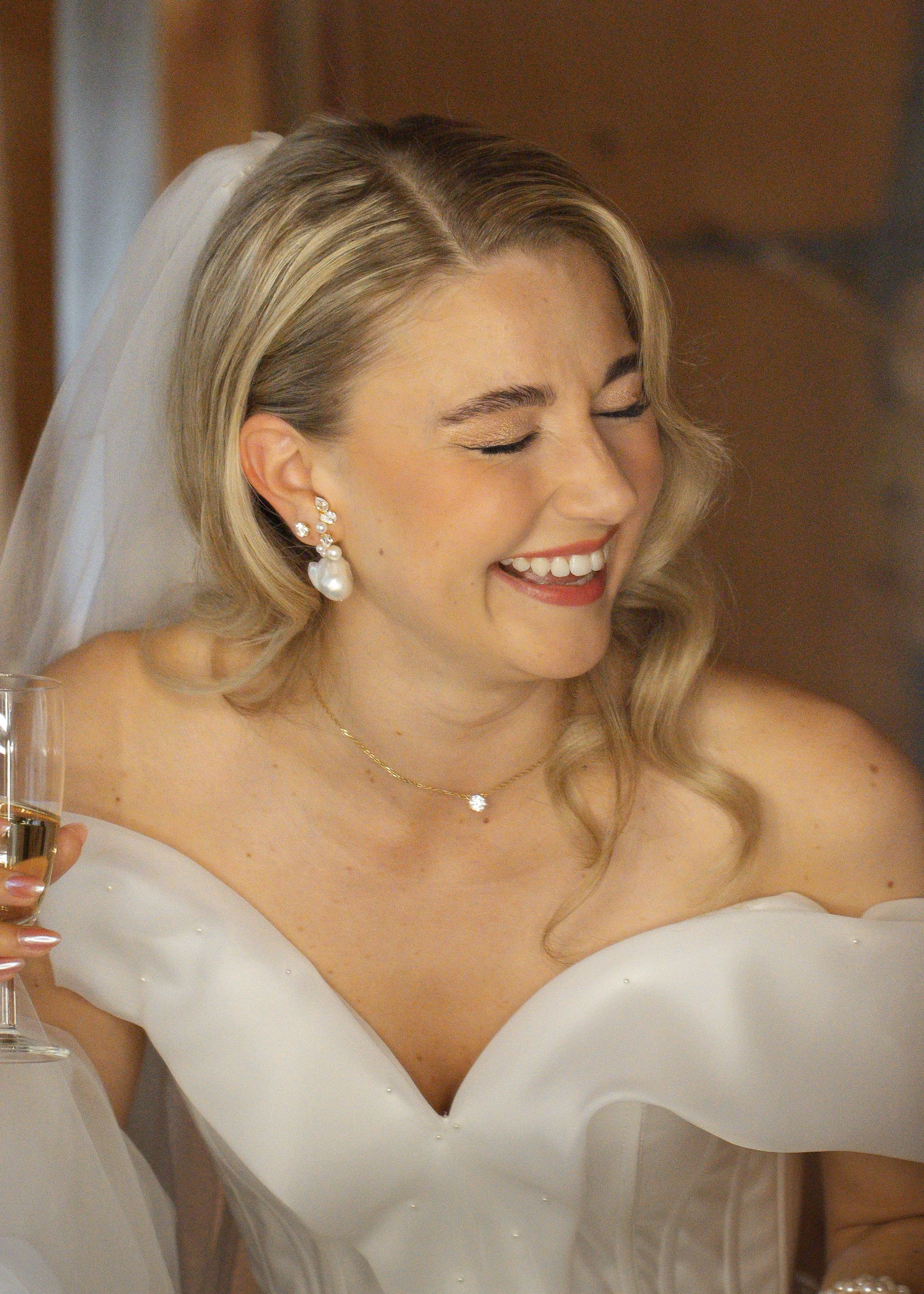 Braut mit weißen Kleid, Perlenohrringen, Halskette und Brautstrauß, lachend bei ihrer Hochzeit.