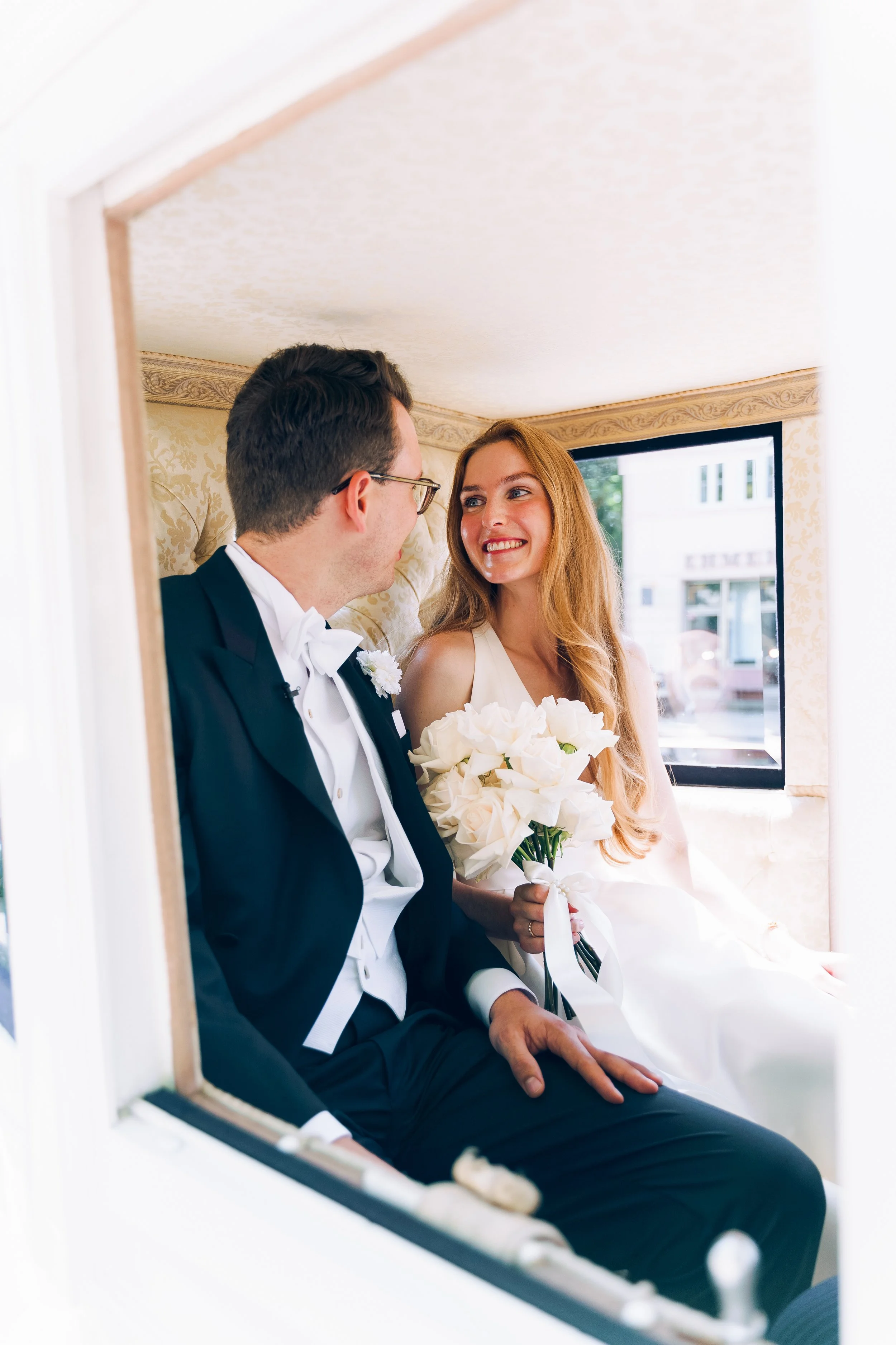 Ein Brautpaar sitzt in einer Kutsche, sie schaut sich liebevoll an, die Braut hält einen weißen Blumenstrauß.