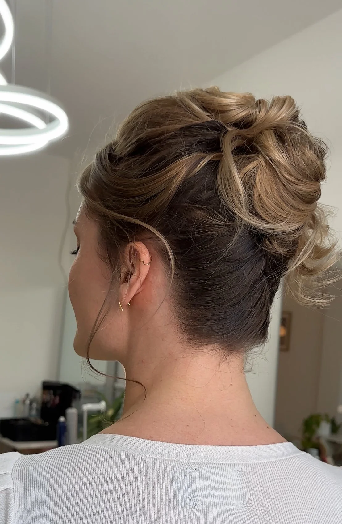 Nahaufnahme eines Damenhaarschnitts mit hochgestecktem, gelocktem Haar, das in einem Salon gestylt wird, sichtbar sind Ohrringe und ein weißes Oberteil.
