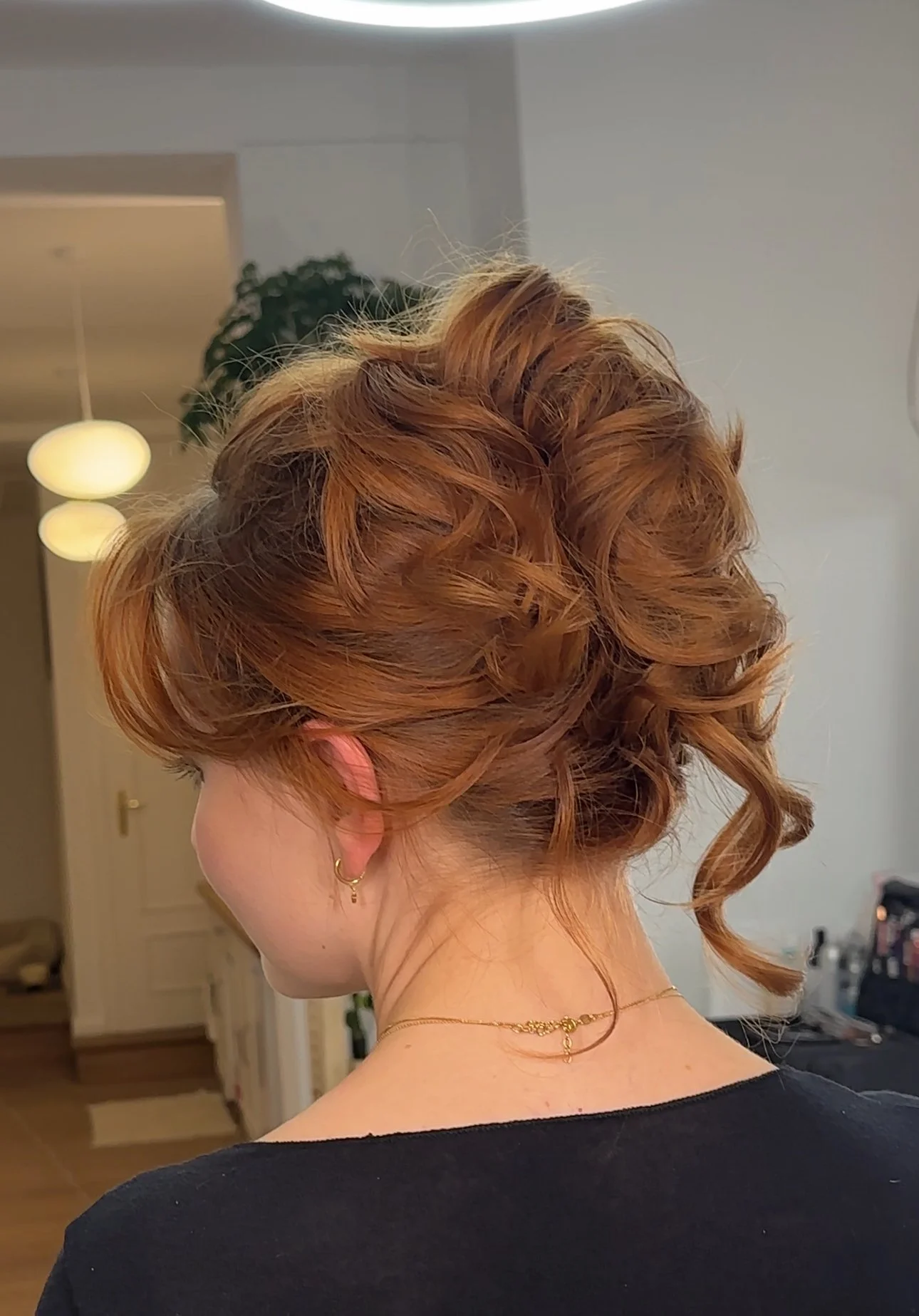 Nebenaufnahme eines Mädchens mit rot-goldenem, lockigem Haar, das eine elegante Frisur trägt, in einem Raum mit gelben Deckenleuchten und einer weißen Wand, getragenes schwarzes Oberteil, Goldschmuck