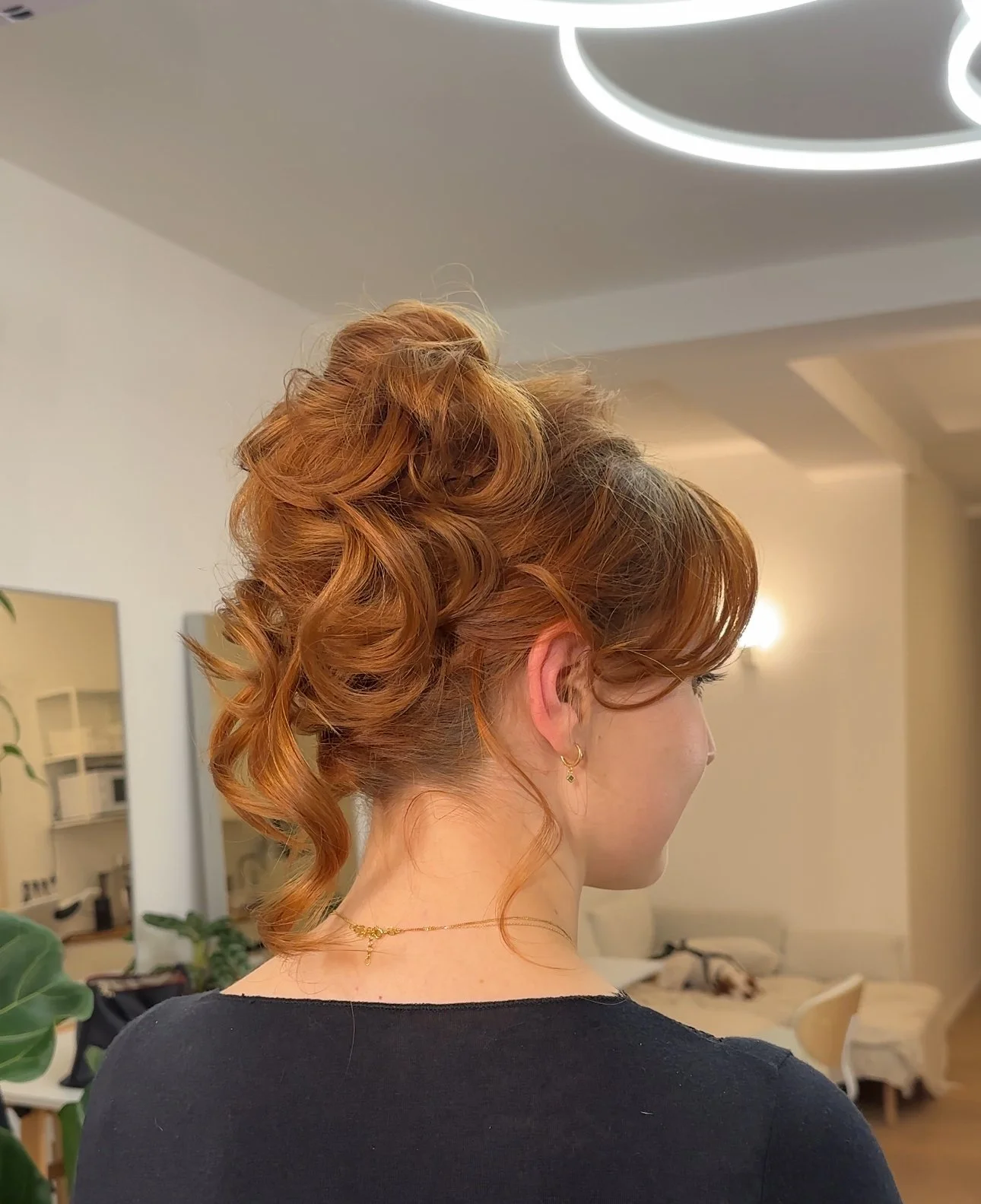 Eine Frau mit roten, lockigen Haaren trägt eine schwarze Kleidung und goldene Schmuckstücke, steht in einem hellen Raum.