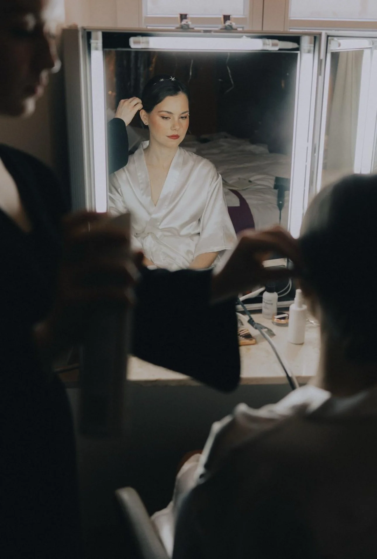 Frau sitzt vor einem Spiegel, während sie sich frisiert, umgeben von Make-up- und Hautpflegeprodukten, im Hintergrund ein Bett.