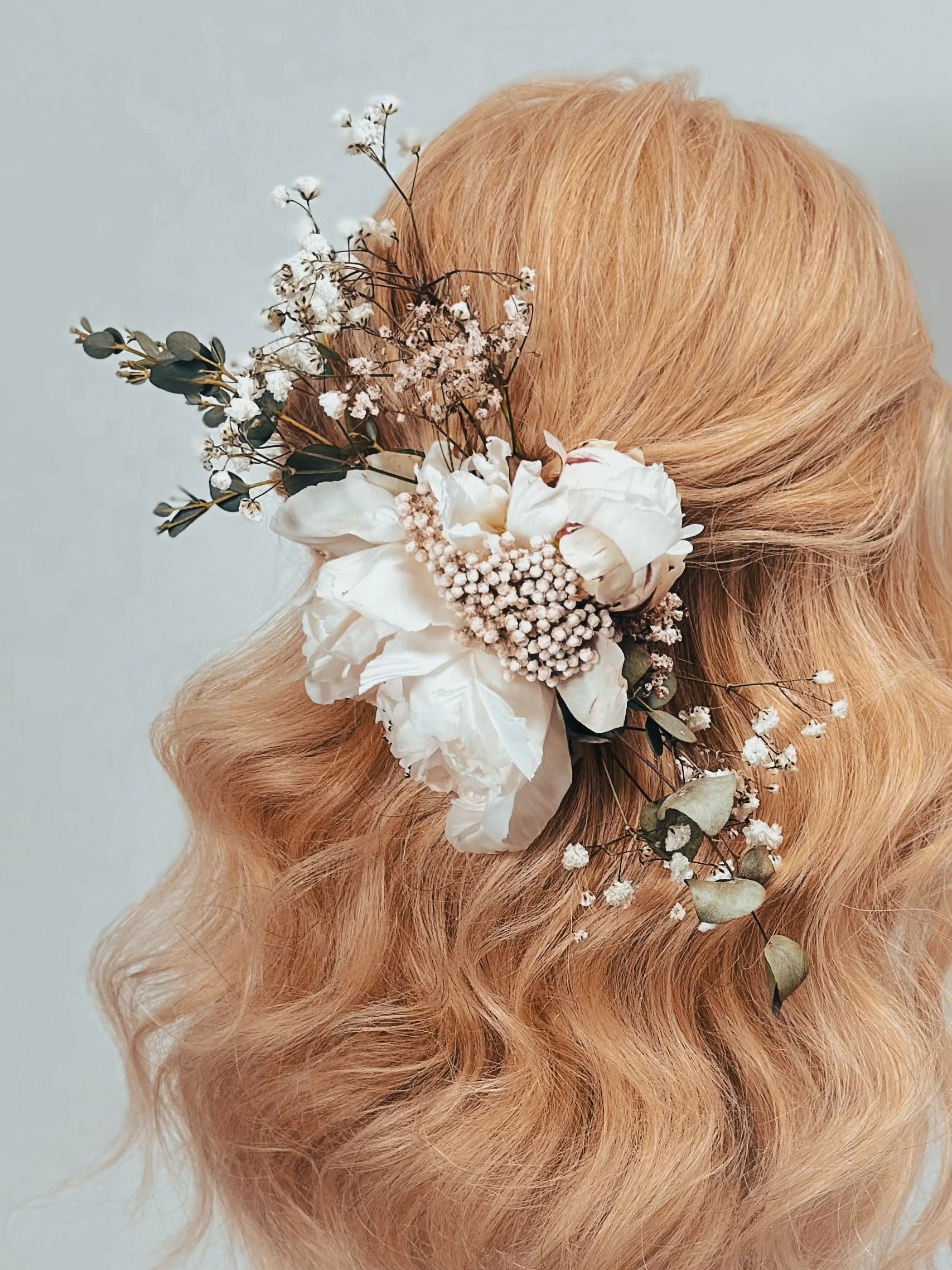Haardekoration mit weißen Blumen, Eukalyptus und kleinen weißen Blüten, die im Haar einer Person mit lockigem, blondem Haar befestigt ist.