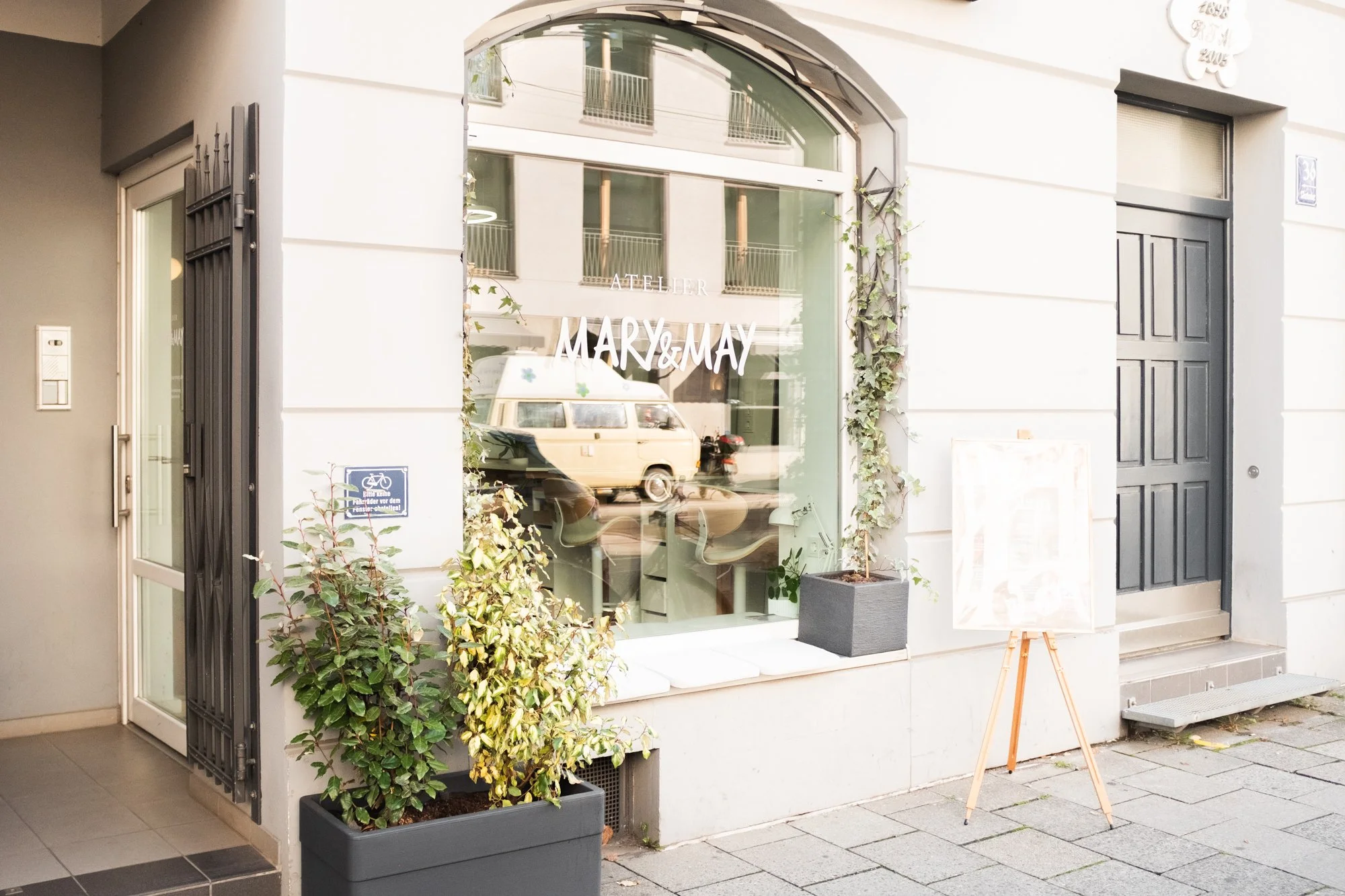 Schaufenster eines Ateliers namens 'Mary & May' mit bepflanzten Fensternischen, einem kleinen weißenboard auf einem Holzständer und einer schwarzen Tür.