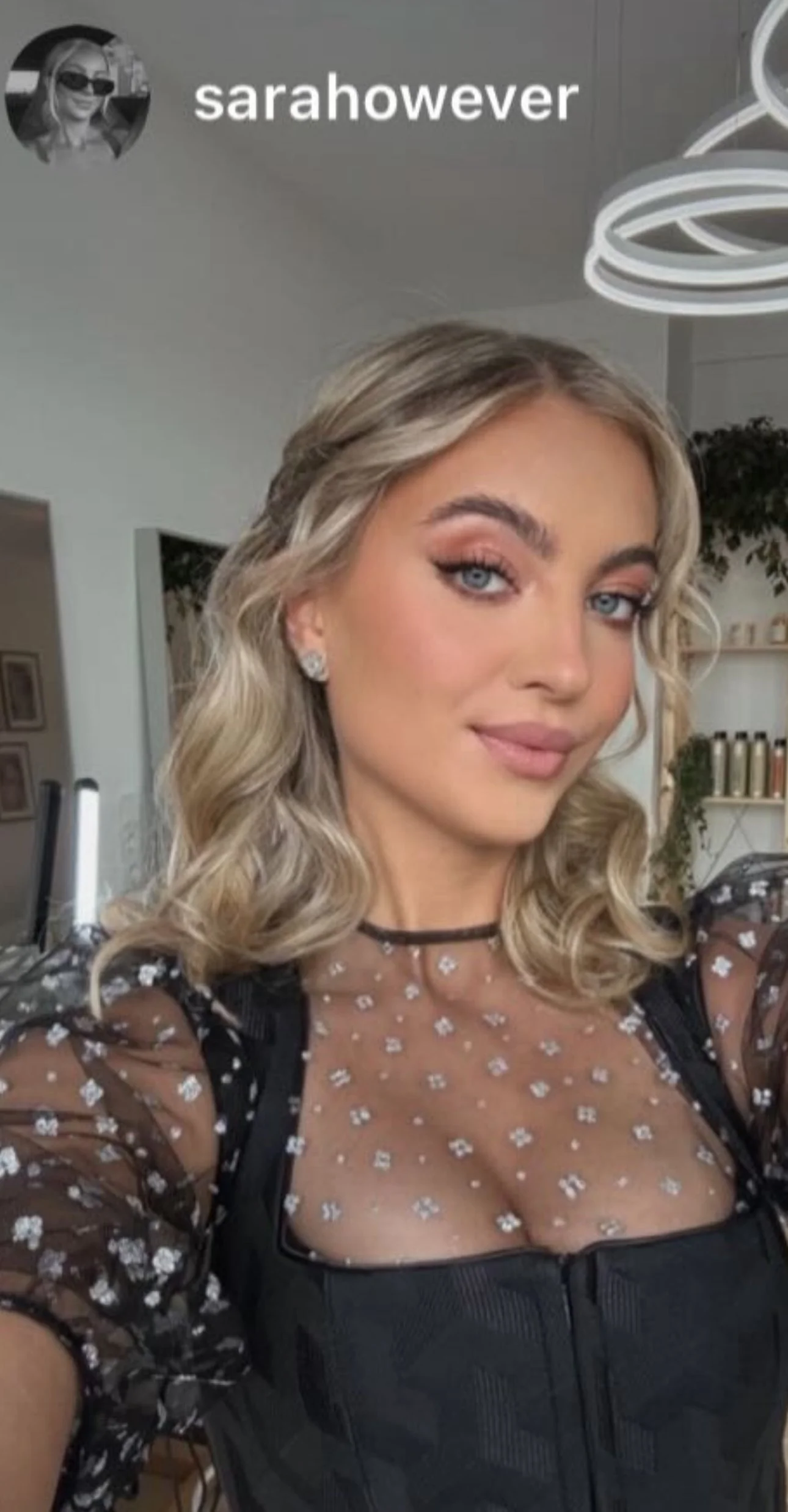 Junge Frau mit blonden, lockigen Haaren, trägt dunklen, durchsichtigen Top mit weißen Blumenmustern, Schmuck, in moderner Inneneinrichtung.