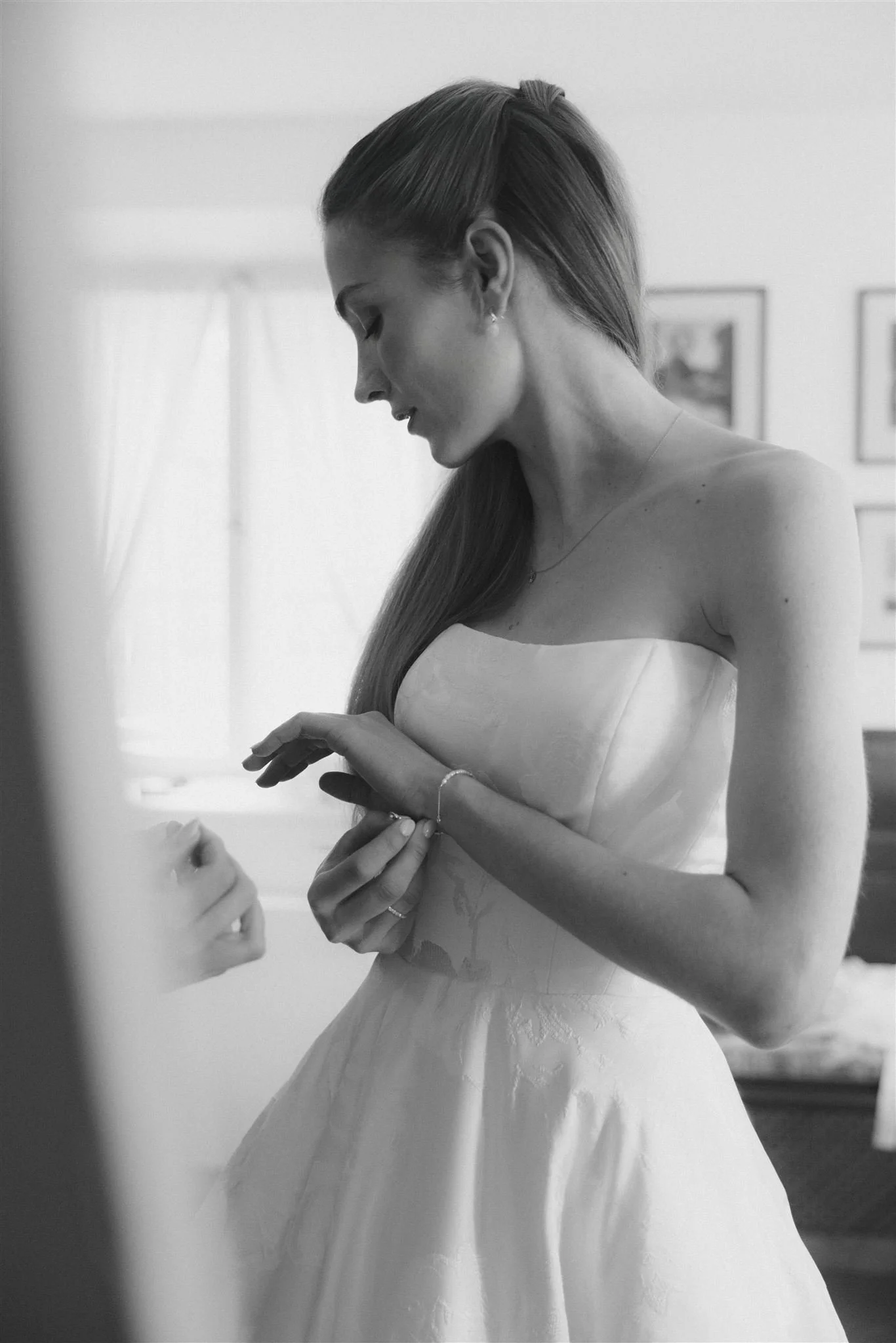 Eine junge Frau in einem Hochzeitskleid schaut nach unten, während sie sich eine Armbanduhr anlegt. Das Bild ist schwarz-weiß, im Hintergrund sind Bilder an der Wand zu sehen.