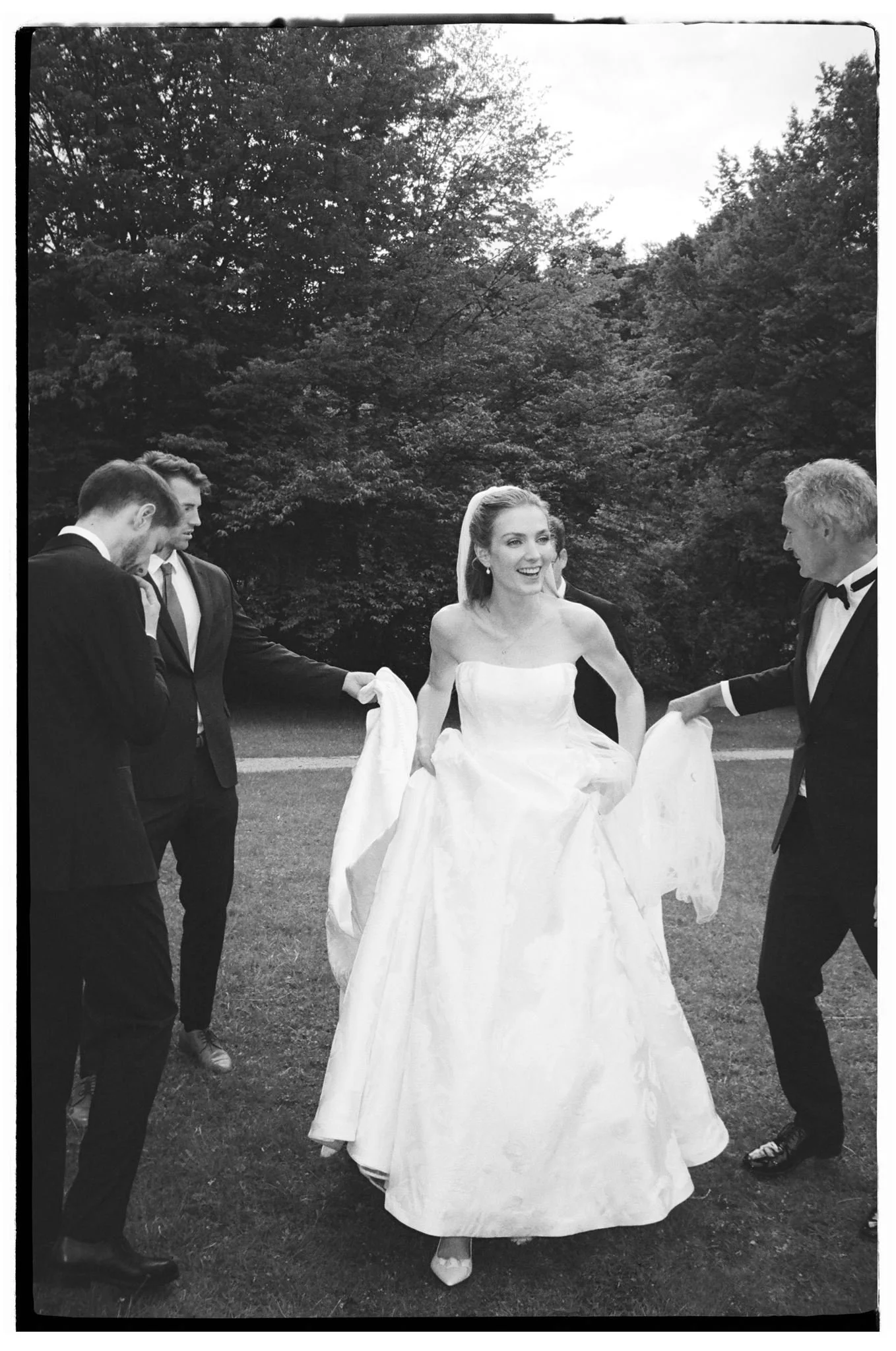 Braut in weißem Brautkleid wird von fünf Männern in dunklen Anzügen umgeben, outdoors im Park.