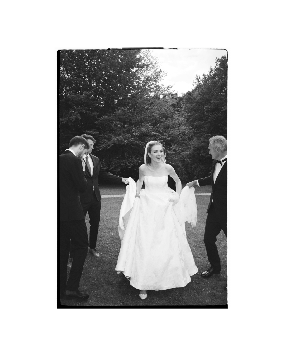 Schwarzweißfoto einer glücklichen Braut in Hochzeitskleid, umgeben von Männern in Anzügen, im Freien mit Bäumen im Hintergrund.