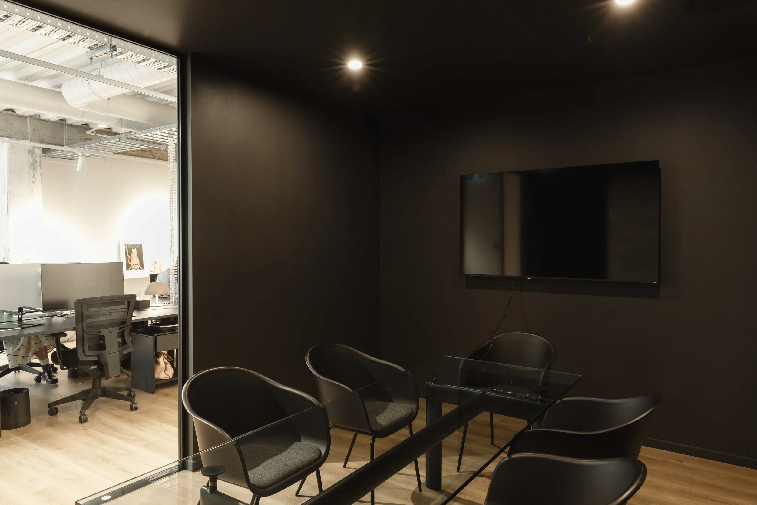 Office conference designed by Zoelen Architects with black chairs, a wall-mounted TV, and a darker wall. Adjacent bright office space visible through a glass wall with desks and chairs, exposed ductwork, and a ceiling.