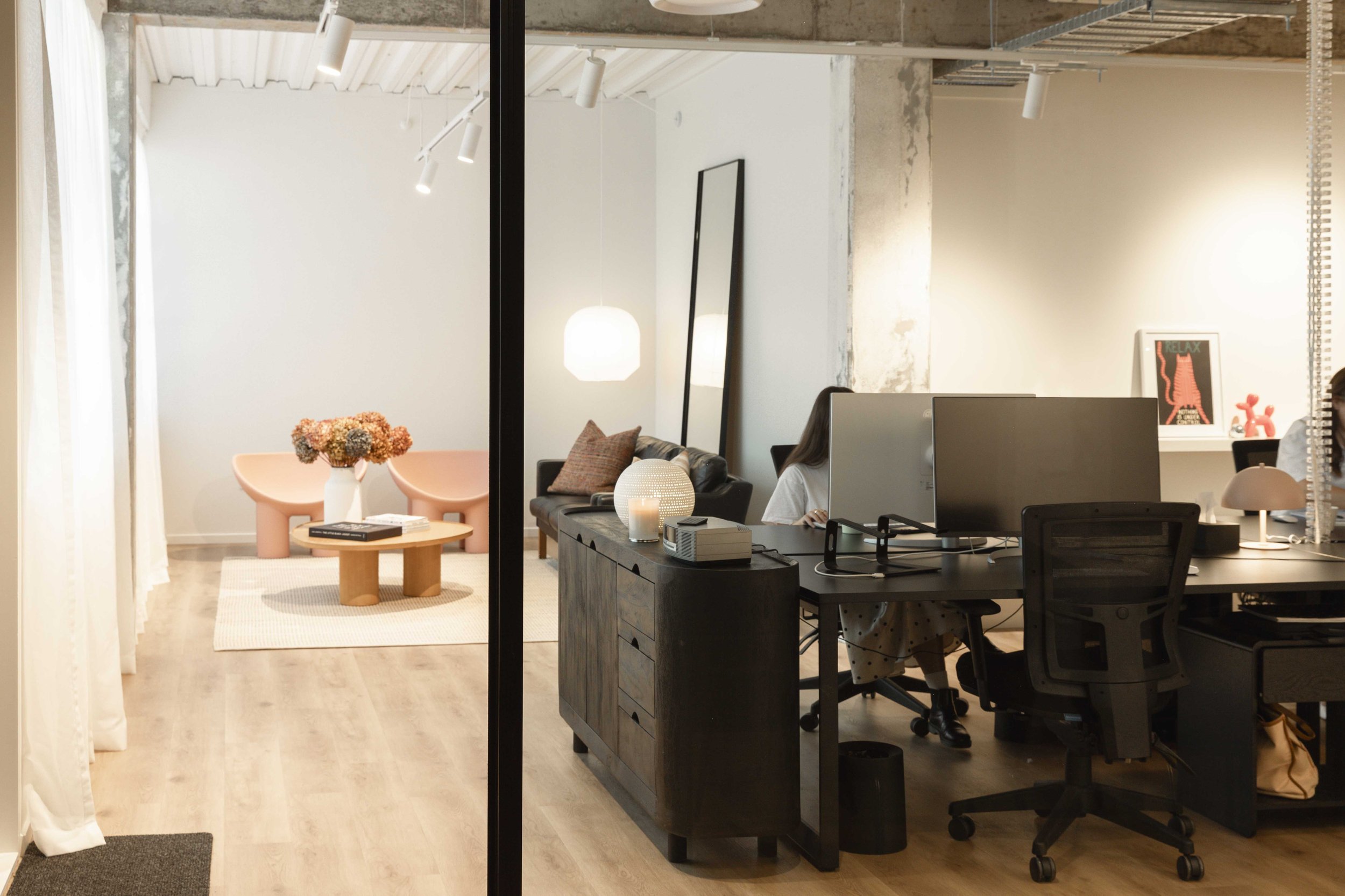 View of a modern office space seen through a glass partition. The office features a wooden floor, a pink table with a flower vase, a cozy seating area with cushions, and desks with computer monitors. The decor includes a white hanging lamp, framed art, and a decorative red dog figure.