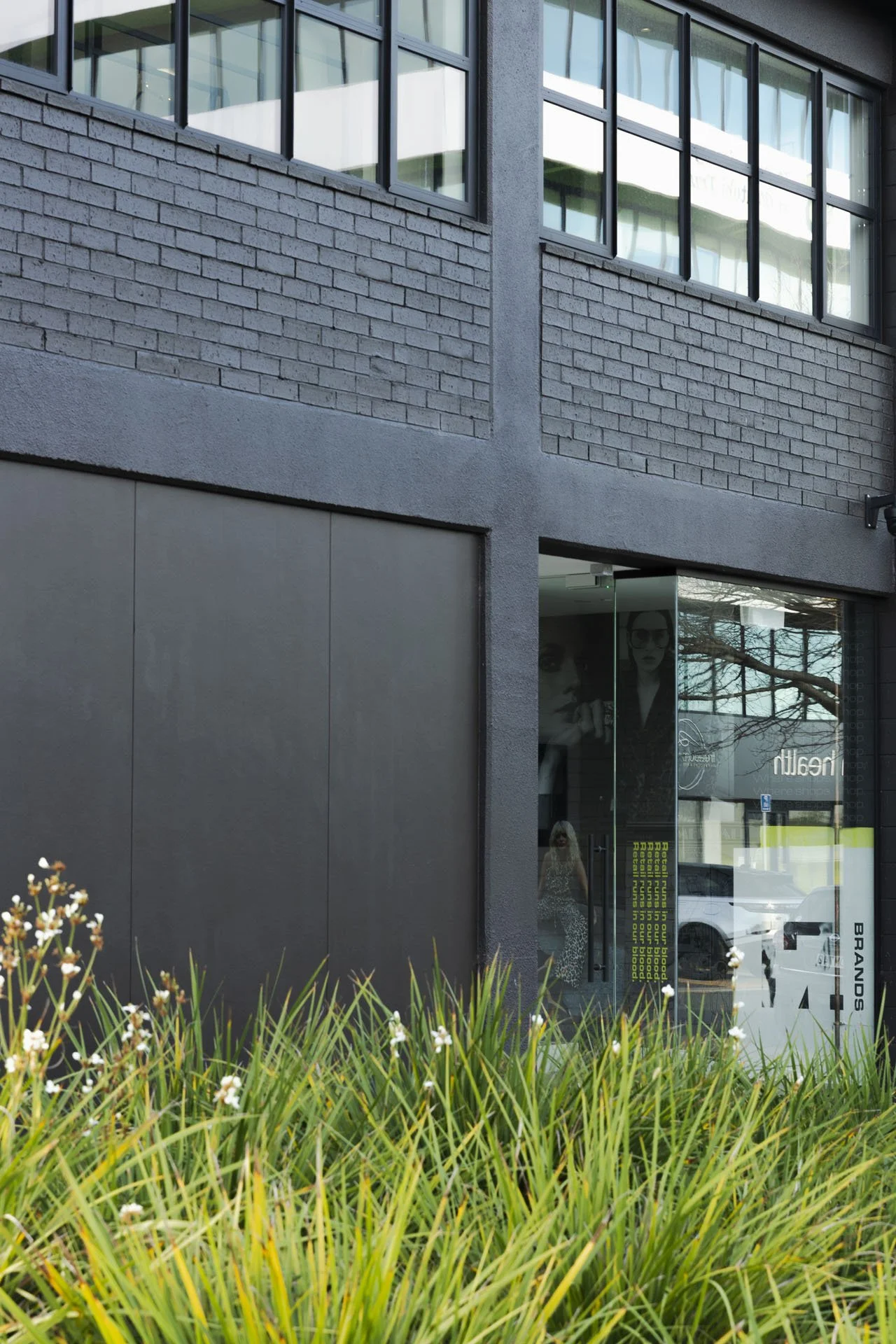 Facade of a modern building with large glass windows, brick and concrete exterior, front entrance with glass door, and some greenery in the foreground.