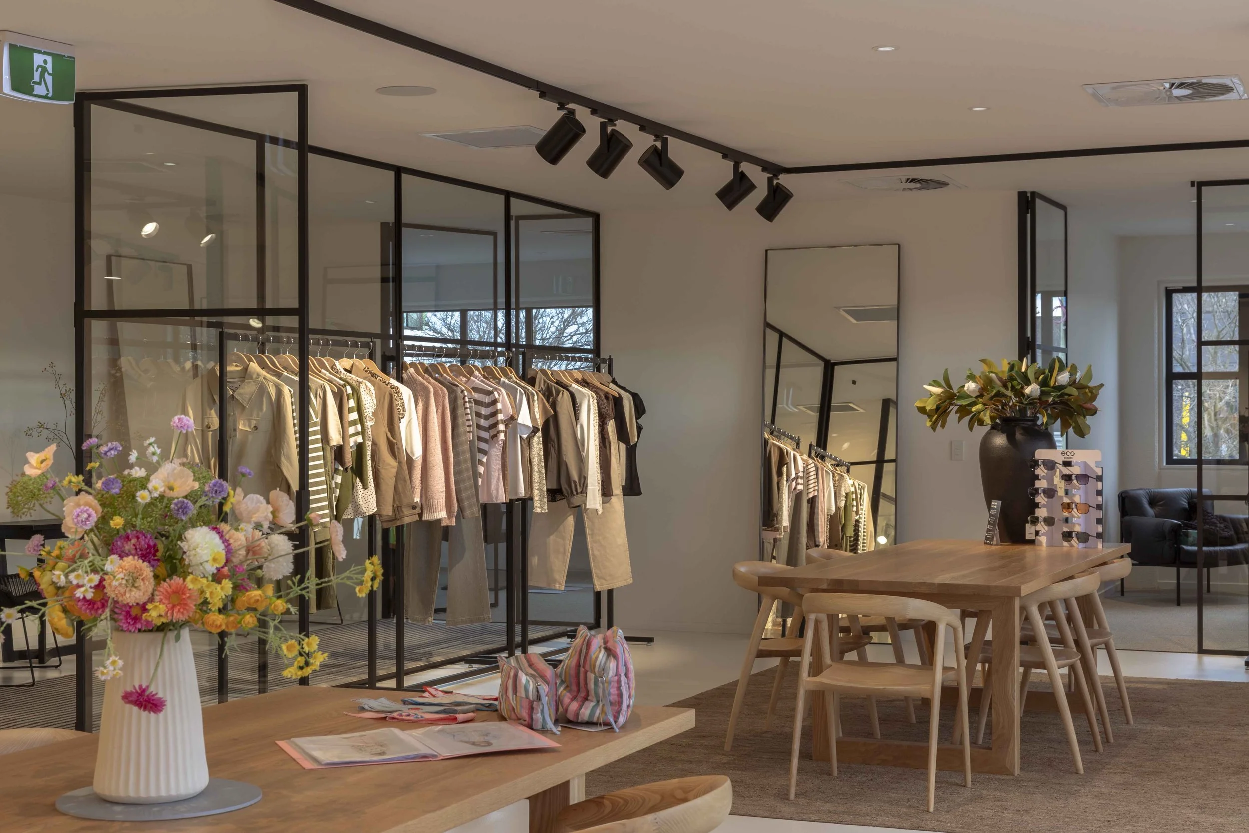 A clothing store with racks of clothing, a large vase with colorful flowers on a wooden table, and a display of sunglasses, in a well-lit modern interior.