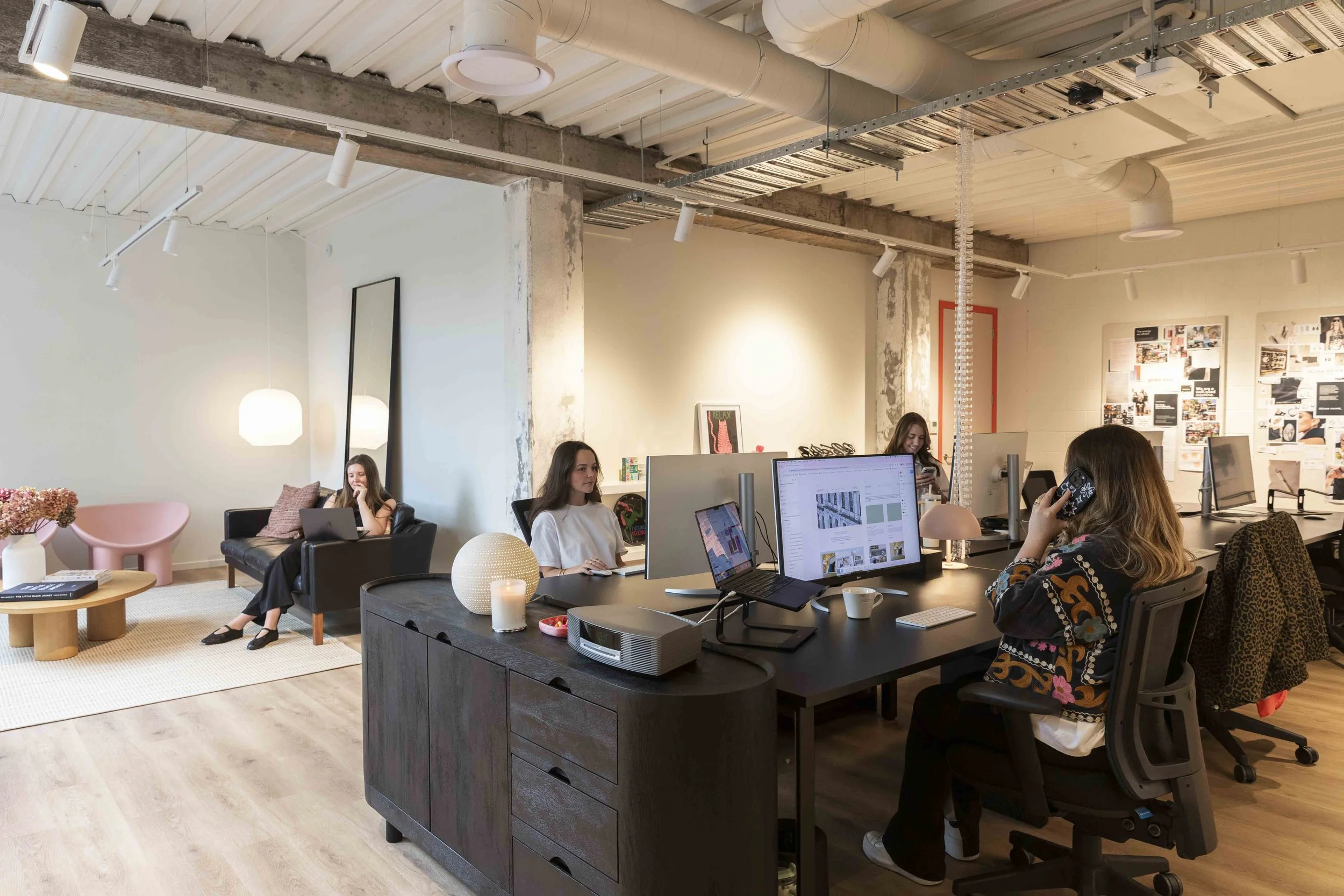 A modern, open-plan office with several women working at desks and a small lounge area with two women sitting and chatting. The office has an industrial ceiling with exposed pipes and ducts, and the walls are decorated with artwork and posters. There are computers, laptops, and office supplies on the desks.