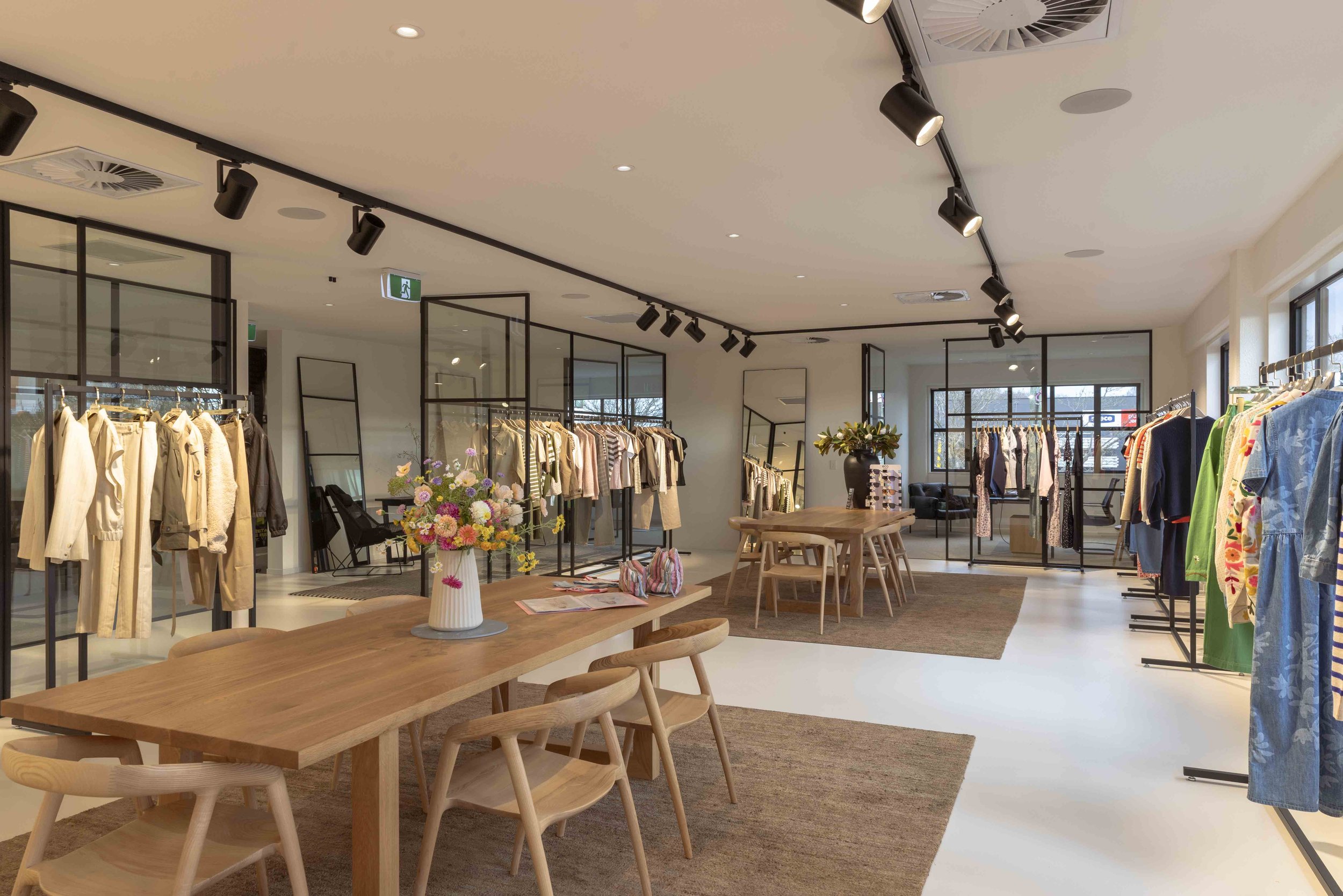 Inside a clothing store with racks of clothes, a wooden table with flowers in a vase, and large windows letting in natural light.