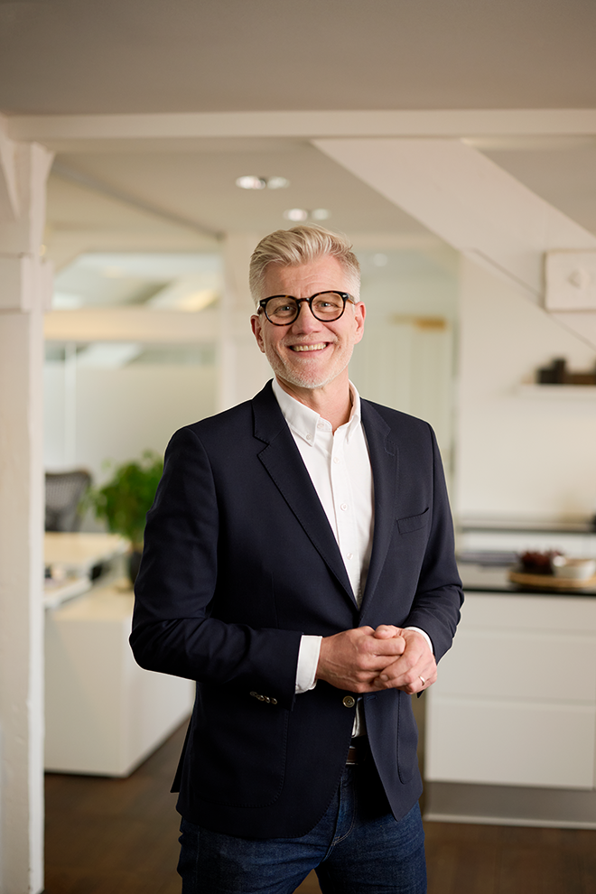 En glad mand med brunt hår, grått skæg og sorte briller, iført en sort blazer og hvid skjorte, står indendørs i et moderne hjem.