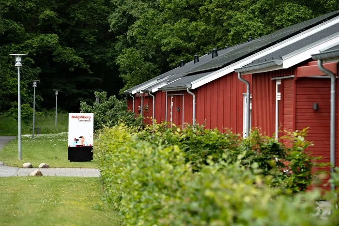 Rød træbungalow med sort tag, beliggende langs en sti med lave buske foran og grønne træer i baggrunden.