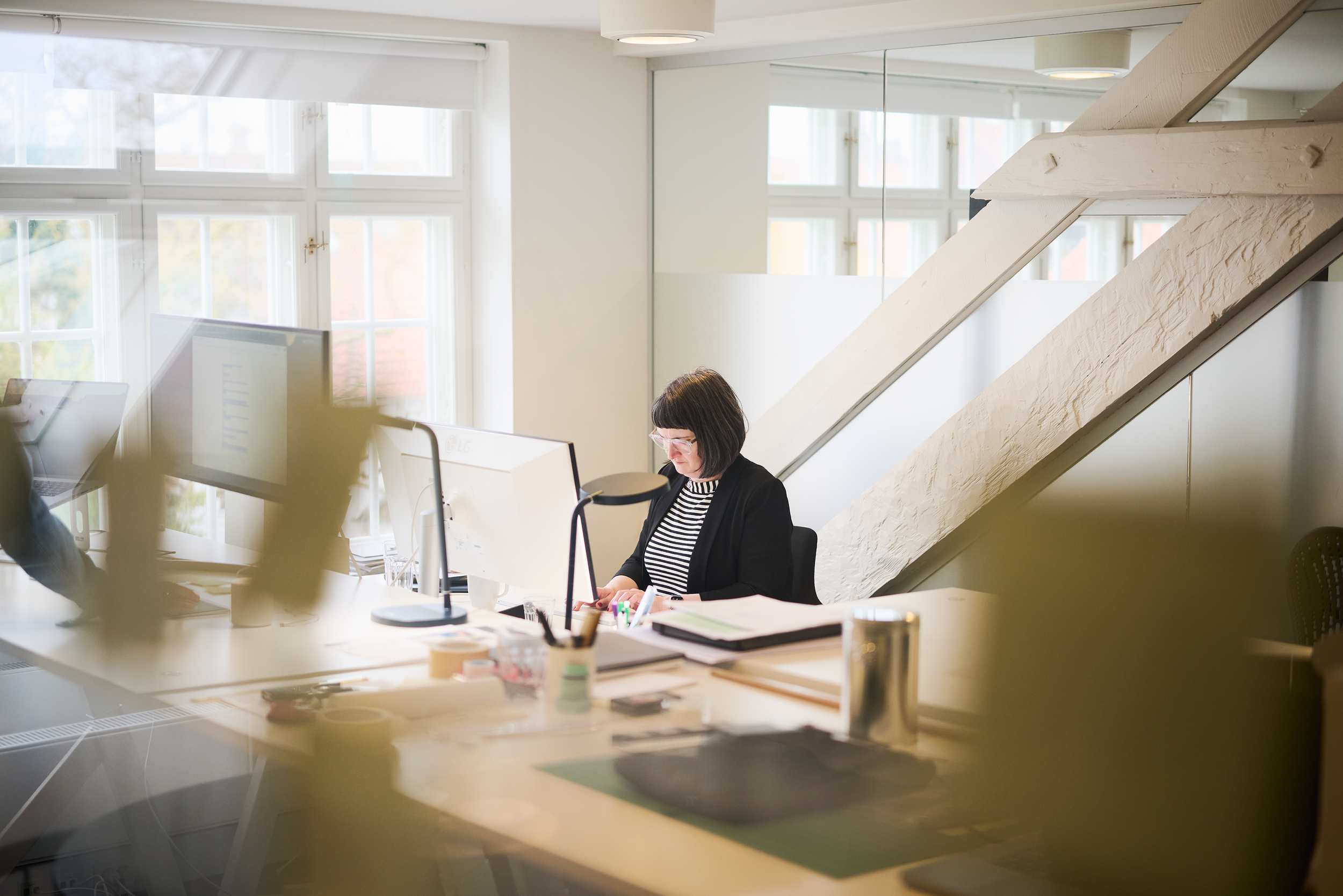 En kvinde arbejder ved sit skrivebord i et kontor med store vinduer, der lader masser af naturligt lys ind.