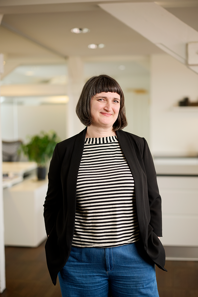 En kvinde med kort mørkt hår og pandehår står i et kontormiljø og smiler.