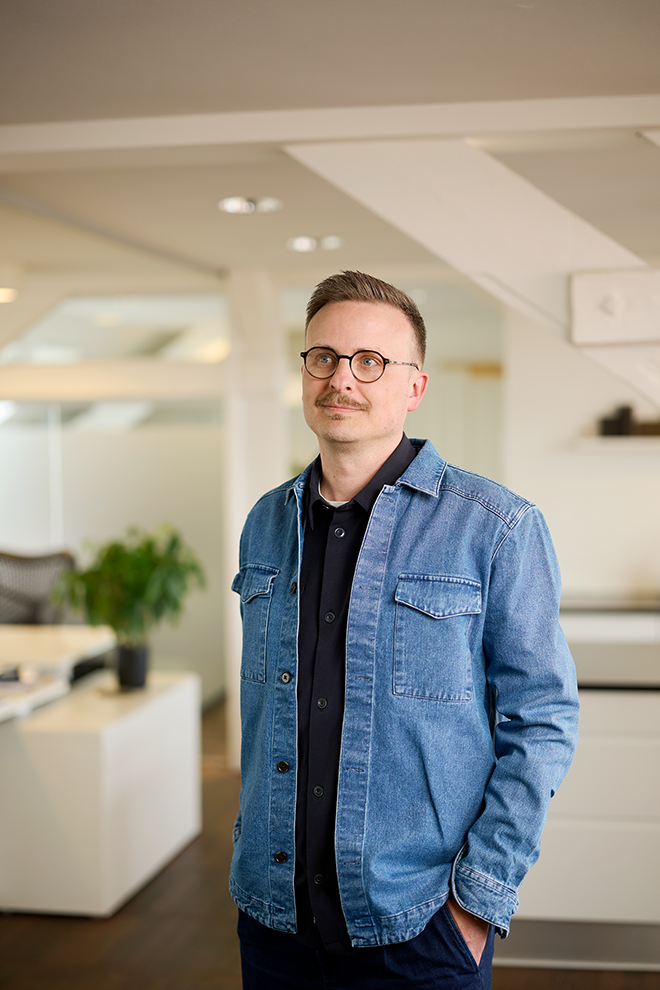 Mand står i moderne kontorlandskab med blød belysning, iført denimjakke og sorte briller.