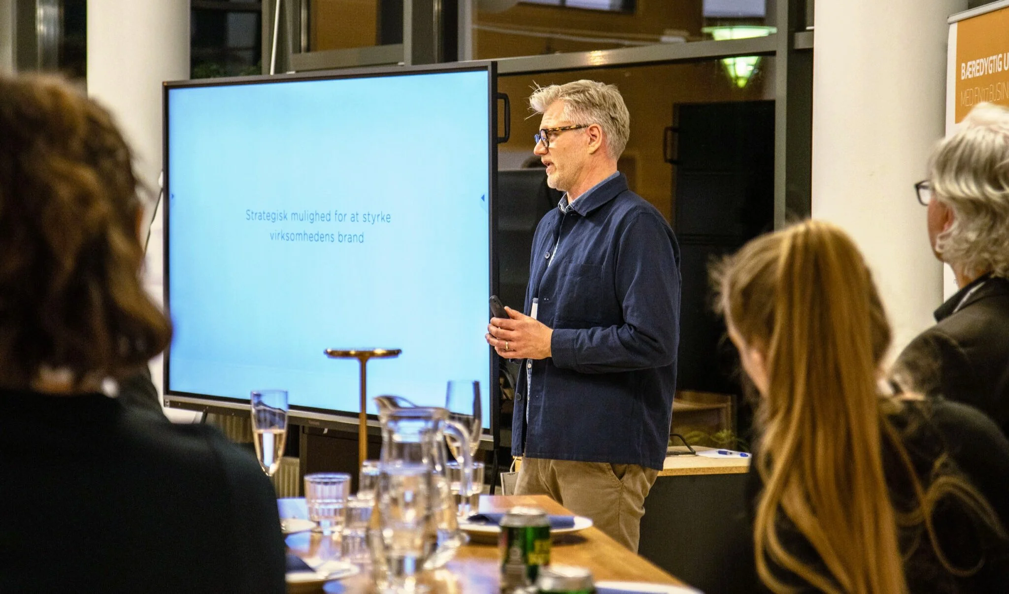 En mand præsenterer foran en stor skærm med teksten "Strategisk mulighed for at styrke virksomhedens brand" under et møde med flere deltagere, der sidder ved et bord med drikke og noter.