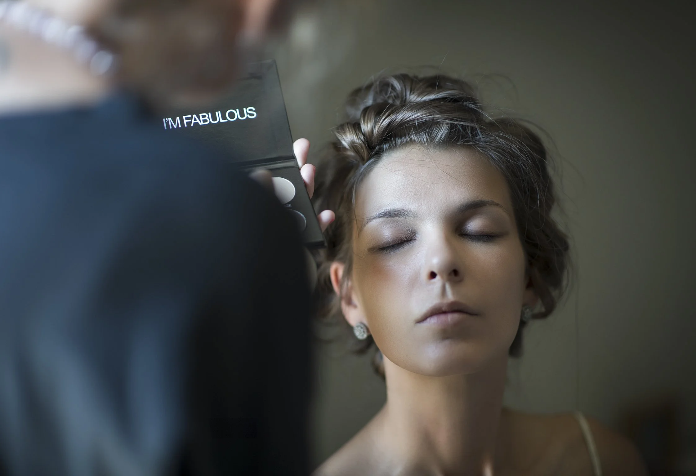 Una donna con gli occhi chiusi viene truccata, mentre una persona tiene una palette di trucco con la scritta "I'm fabulous".