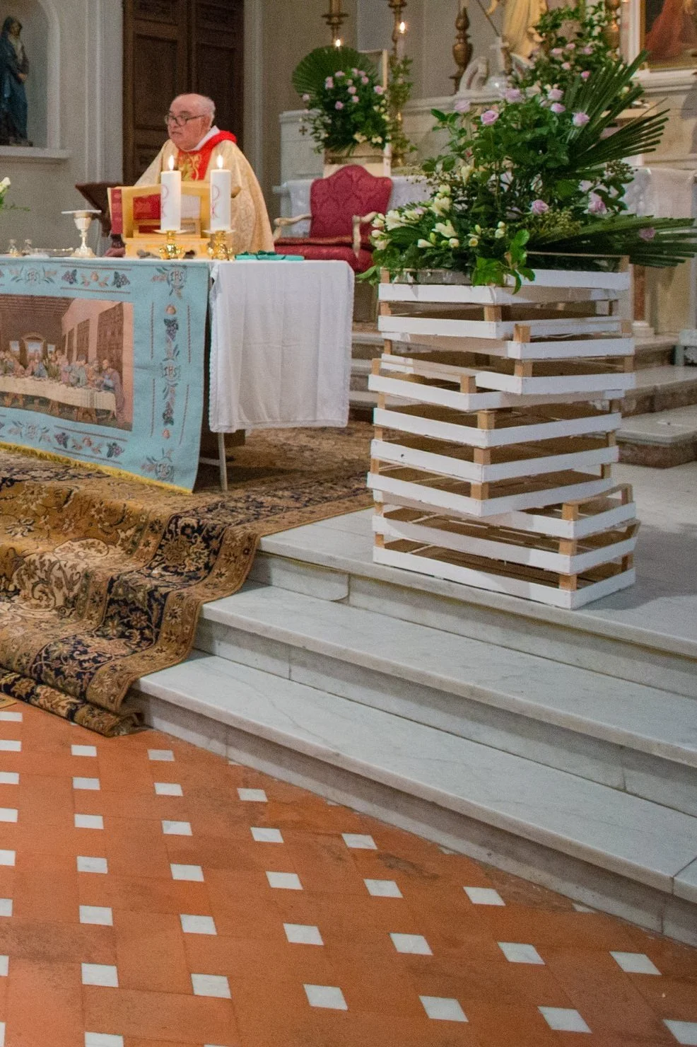 Cerimonia di matrimonio in chiesa, con una coppia di sposi seduti davanti all'altare e un sacerdote che officia il rito. Sullo sfondo, decorazioni floreali e un altare con tovaglia decorata.