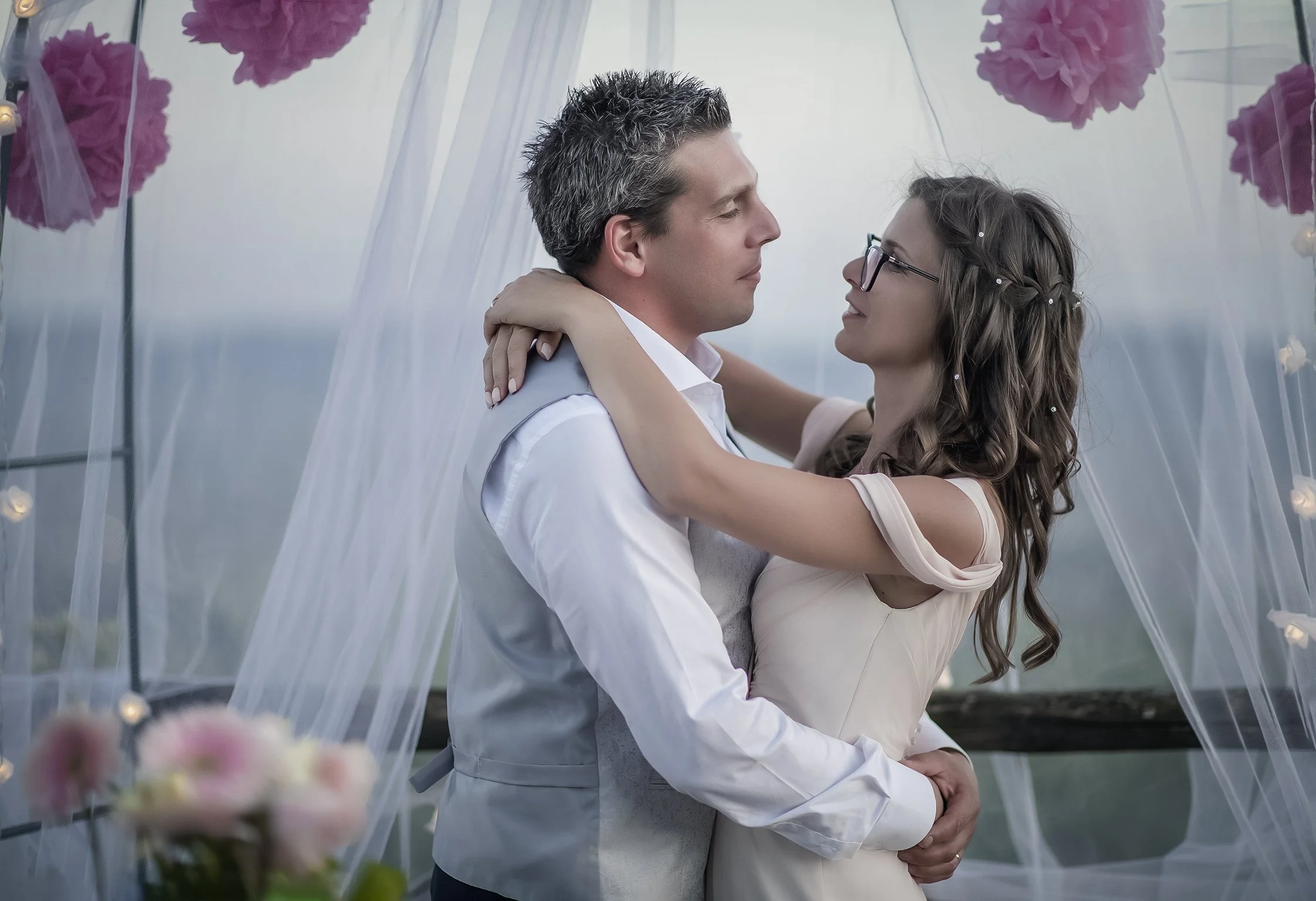 Una coppia abbracciata sotto decorazioni floreali rosa e veli bianchi, probabilmente durante una cerimonia o festa romantica.