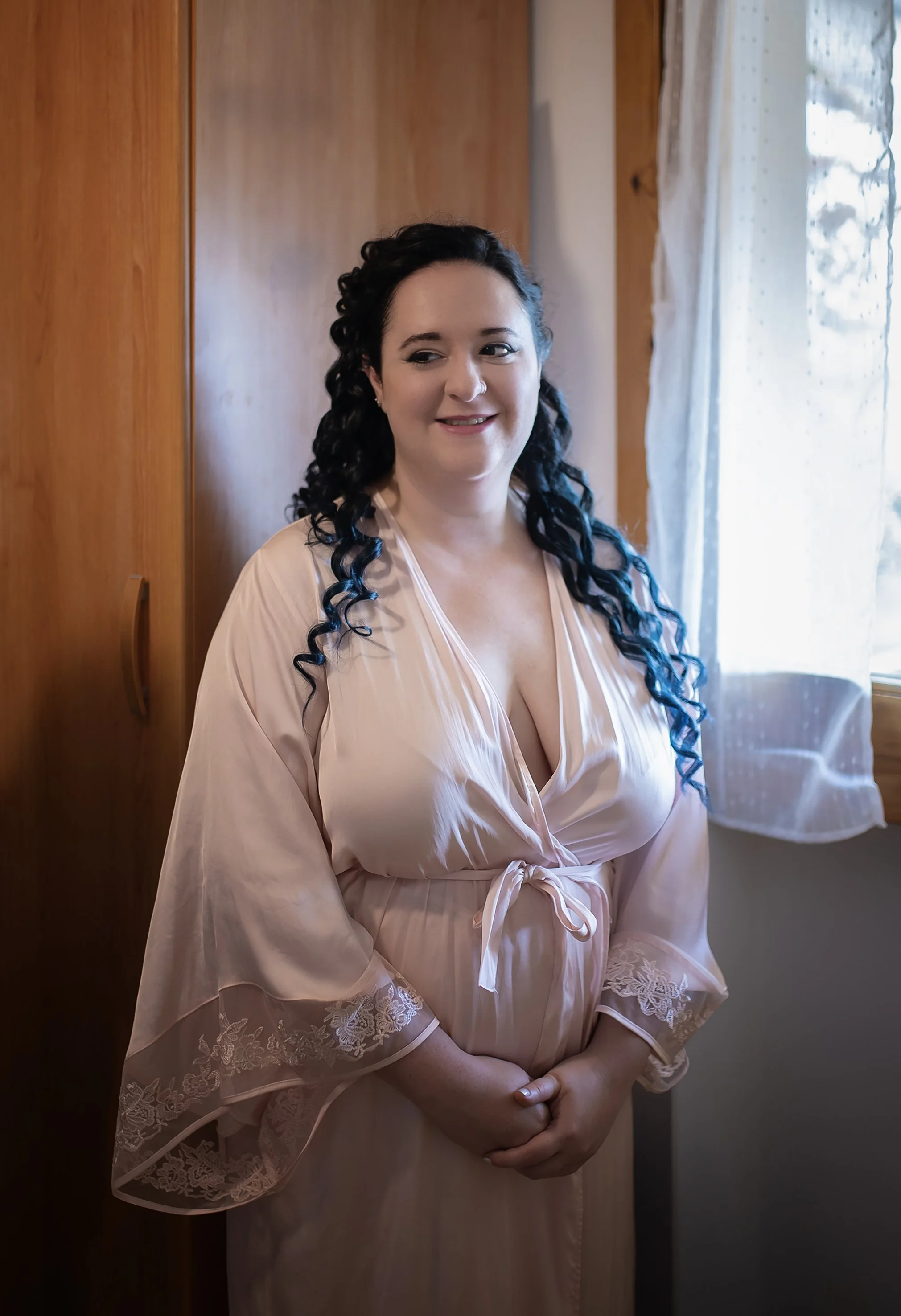 Donna con capelli ricci neri in vestaglia rosa chiara, in piedi accanto a una finestra e un armadio.