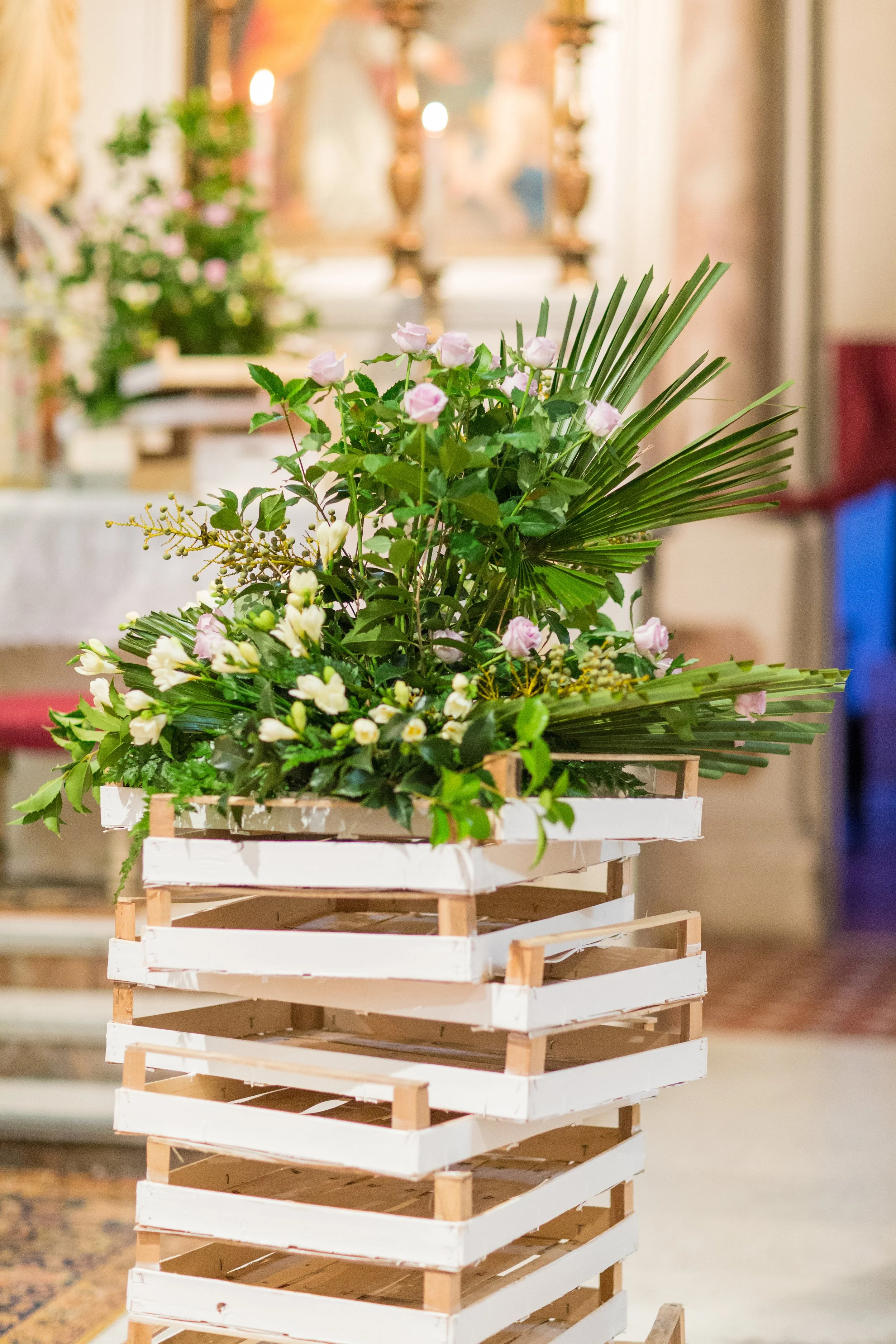 Composizione floreale con rose rosa e foglie di palma in una cassa di legno bianca, interno di una chiesa.