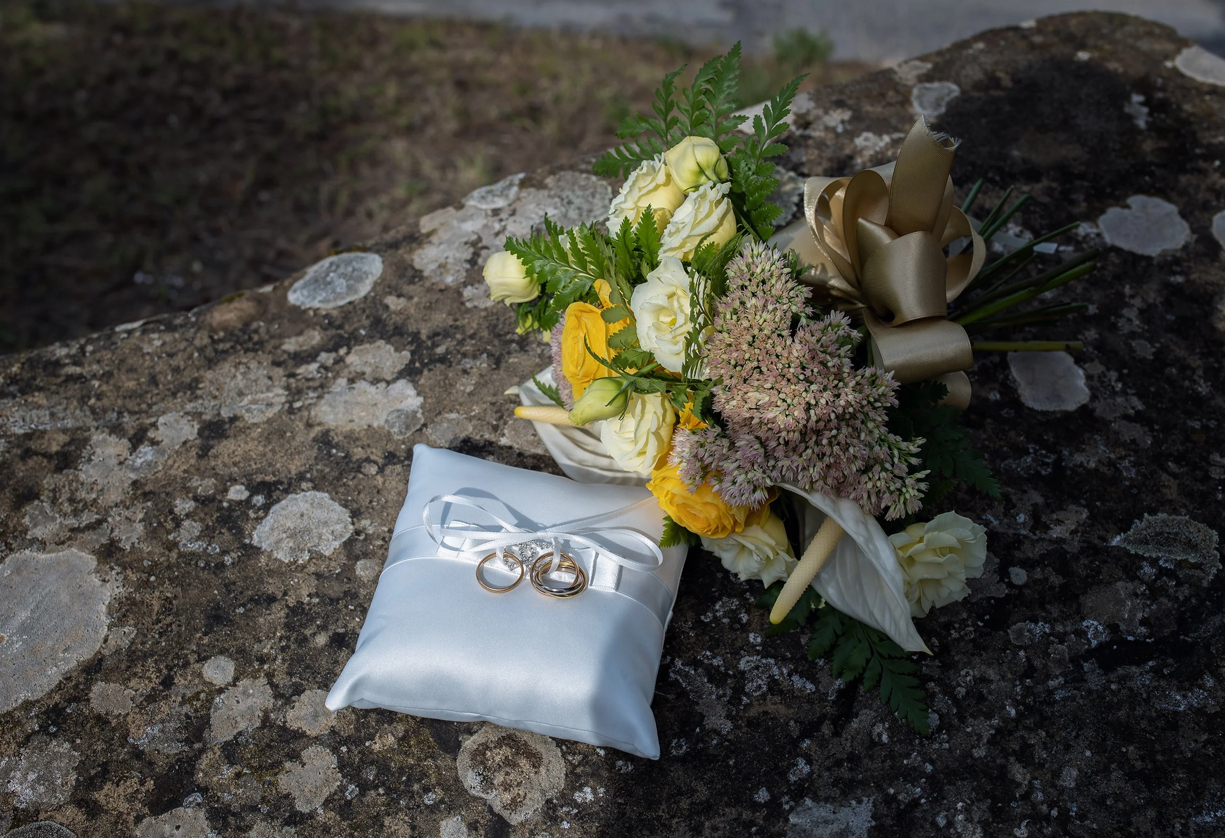Cuscino bianco con fedi nuziali e bouquet di fiori su una superficie di pietra.