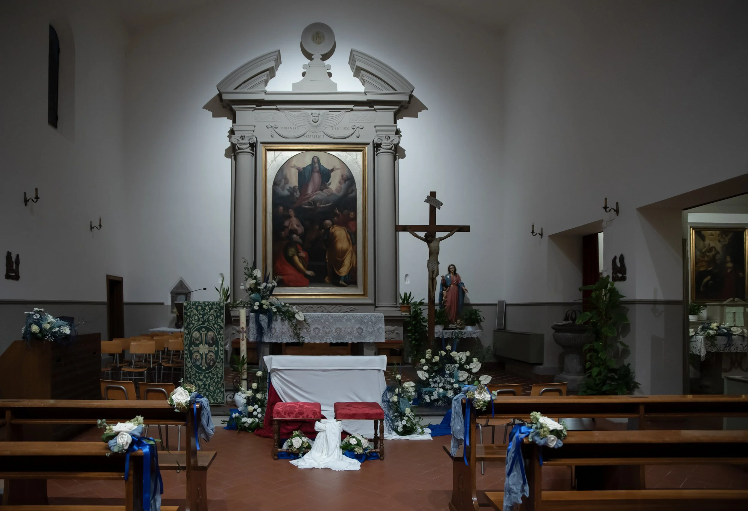 Interno di una chiesa con altare decorato per un matrimonio, crocifisso e statua religiosa, panchine con fiori bianchi e blu.