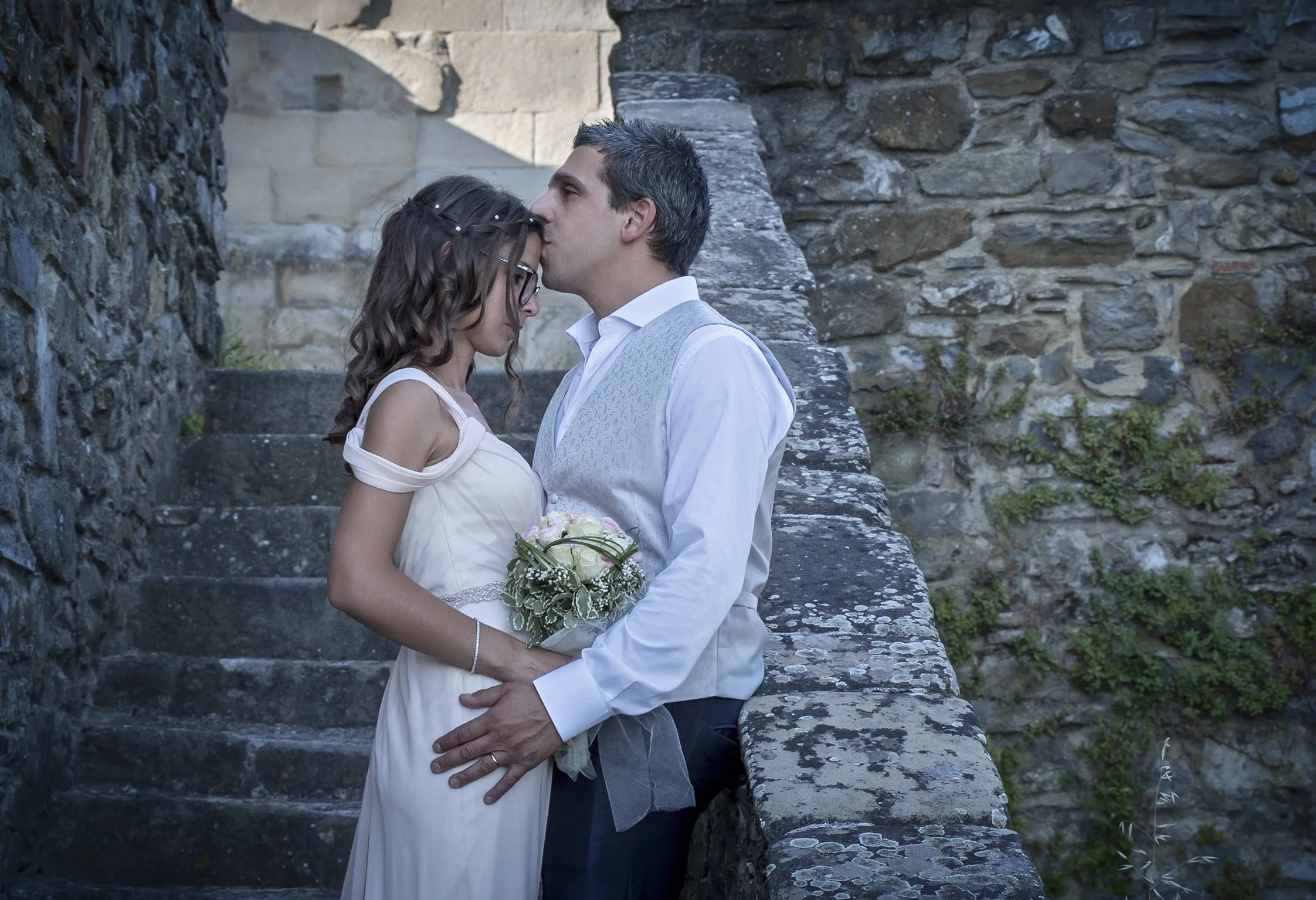 Due persone si abbracciano in abiti da matrimonio accanto a un muro di pietra.