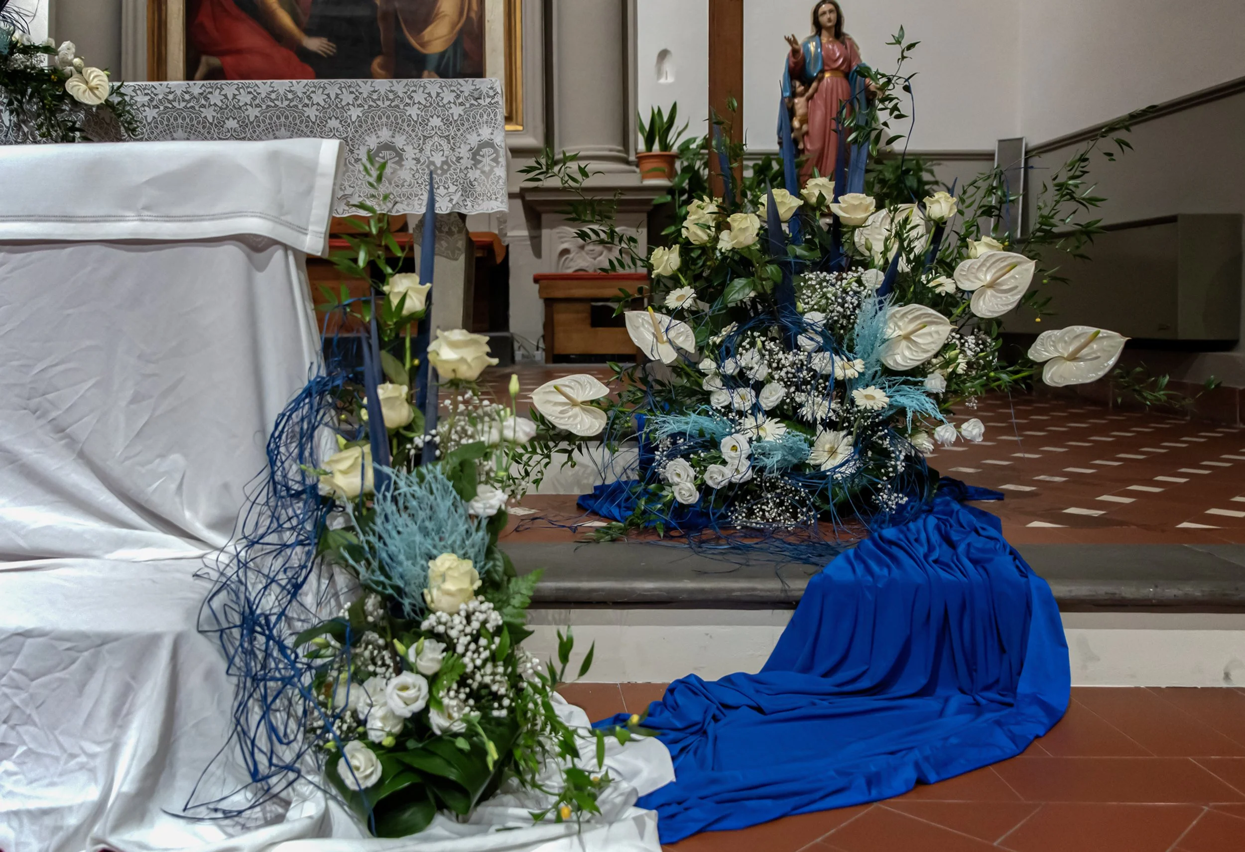 Decorazione floreale con rose e calle bianche su un altare con drappi blu e una statua religiosa sullo sfondo.