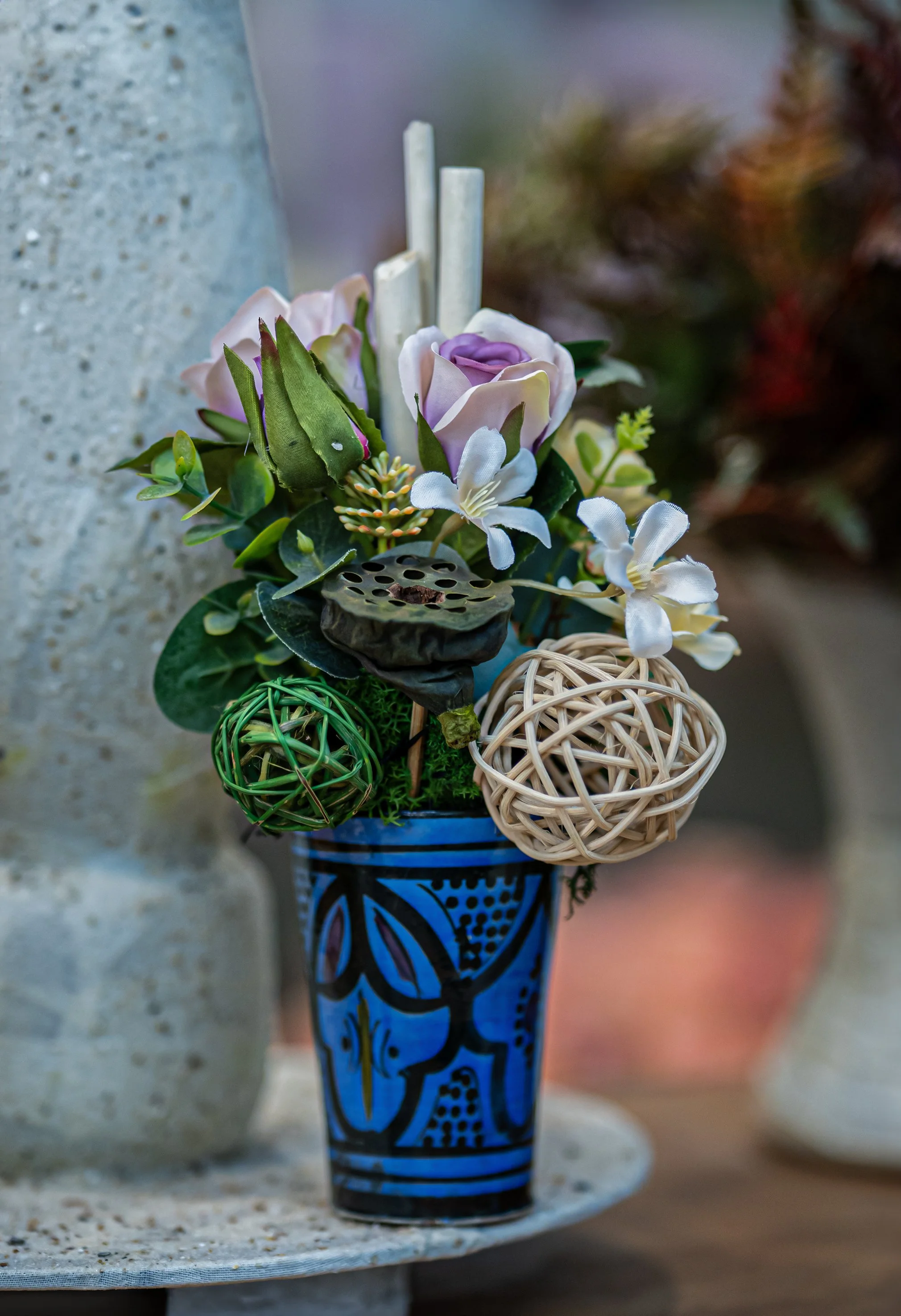 Un vaso blu decorato contiene una composizione floreale con fiori artificiali, foglie verdi e decorazioni sferiche intrecciate.
