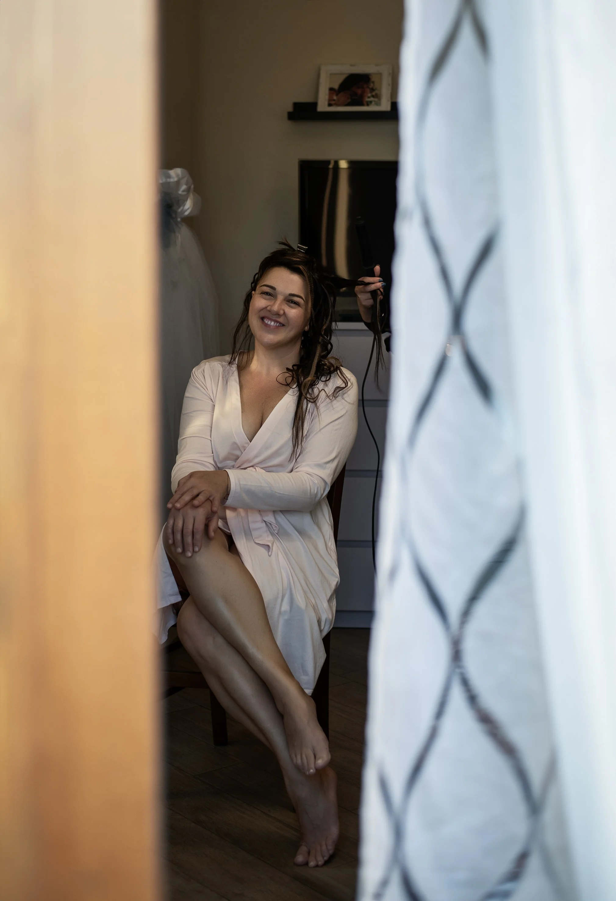 Donna che sorride mentre si prepara per un evento, con i capelli acconciati, indossa un accappatoio e siede su una sedia, con tende ai lati.