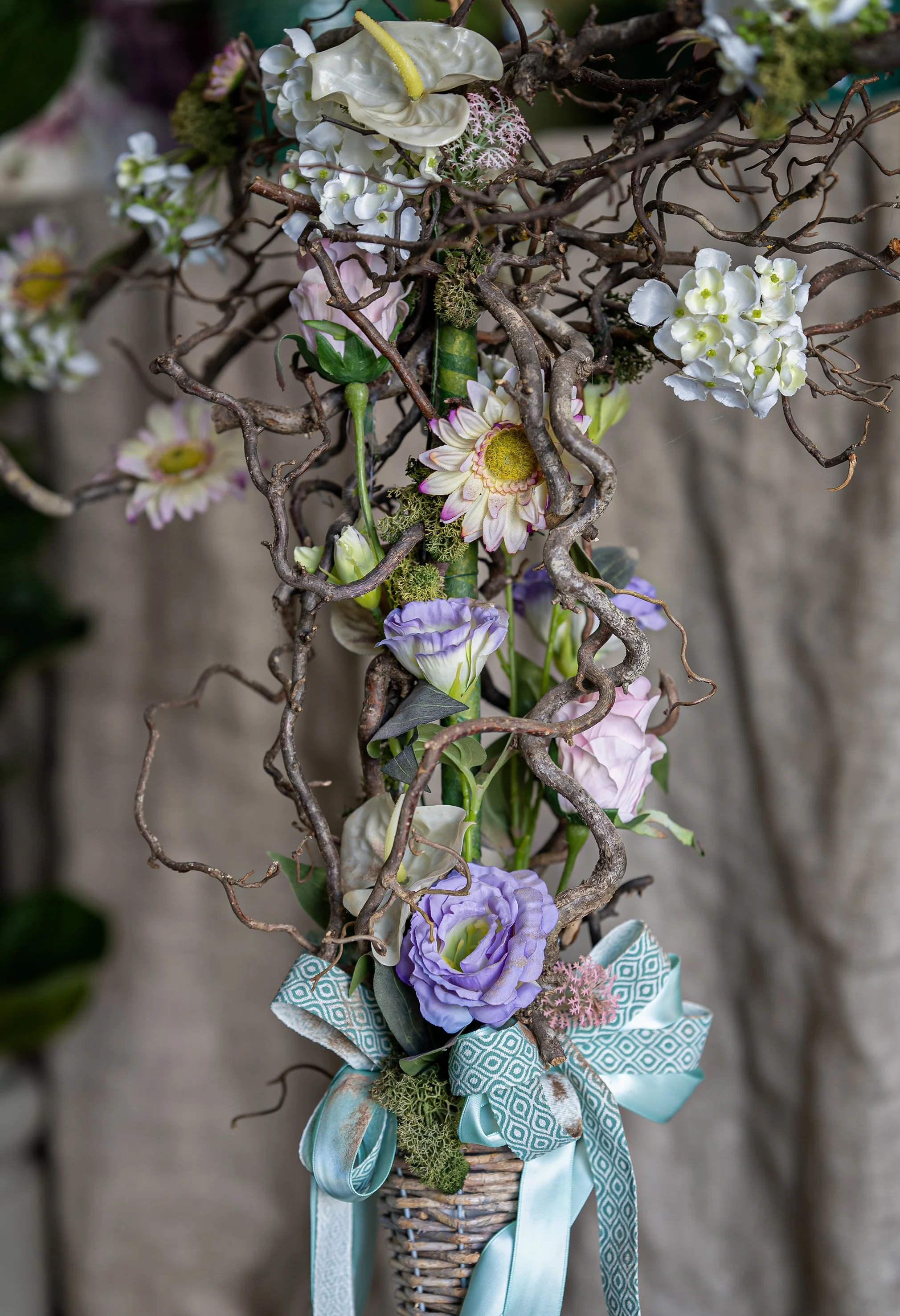 Composizione floreale con fiori bianchi, viola e rosa intrecciati a rami secchi, decorata con nastro azzurro.