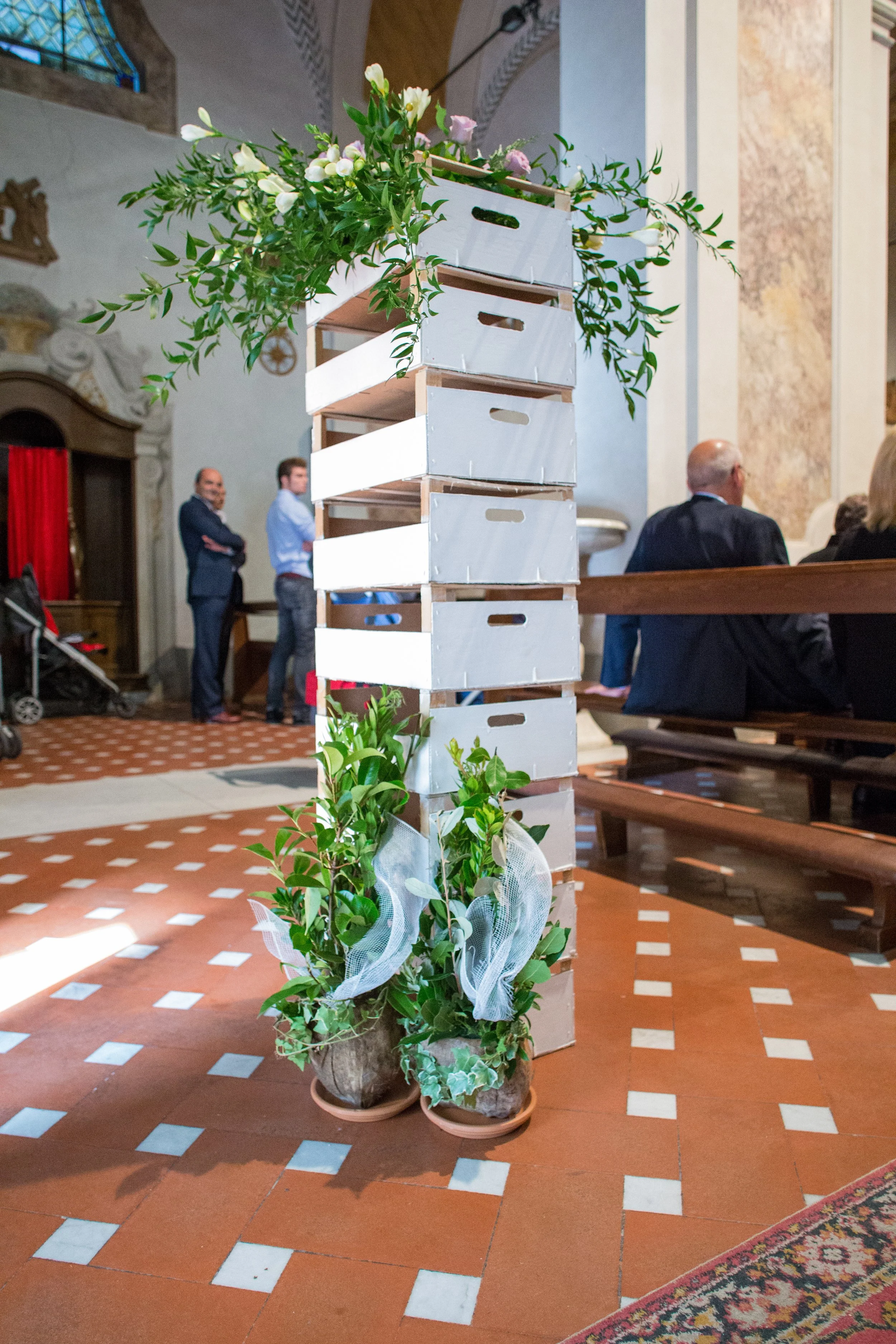Decorazione floreale in chiesa con cassette di legno bianche impilate e vasi di piante alla base.