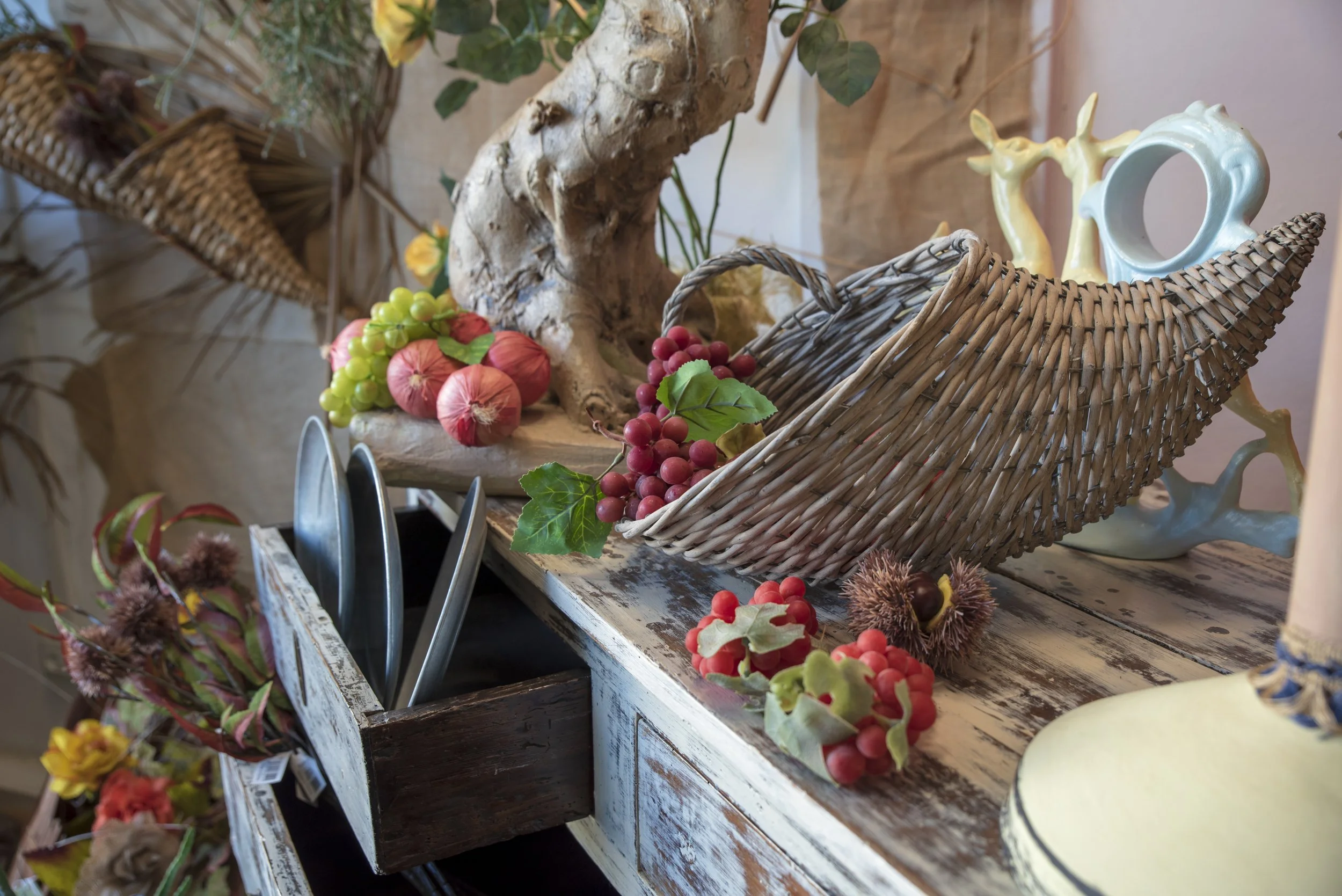 Decorazione autunnale con cesto di vimini, frutta artificiale, foglie verdi e elementi naturali su una superficie di legno.