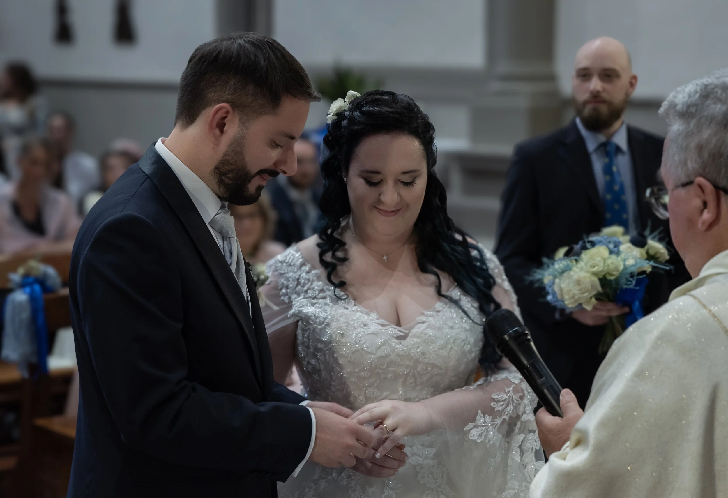 Una coppia si sposa, lo sposo mette l'anello al dito della sposa in chiesa, con un sacerdote e testimone presenti.