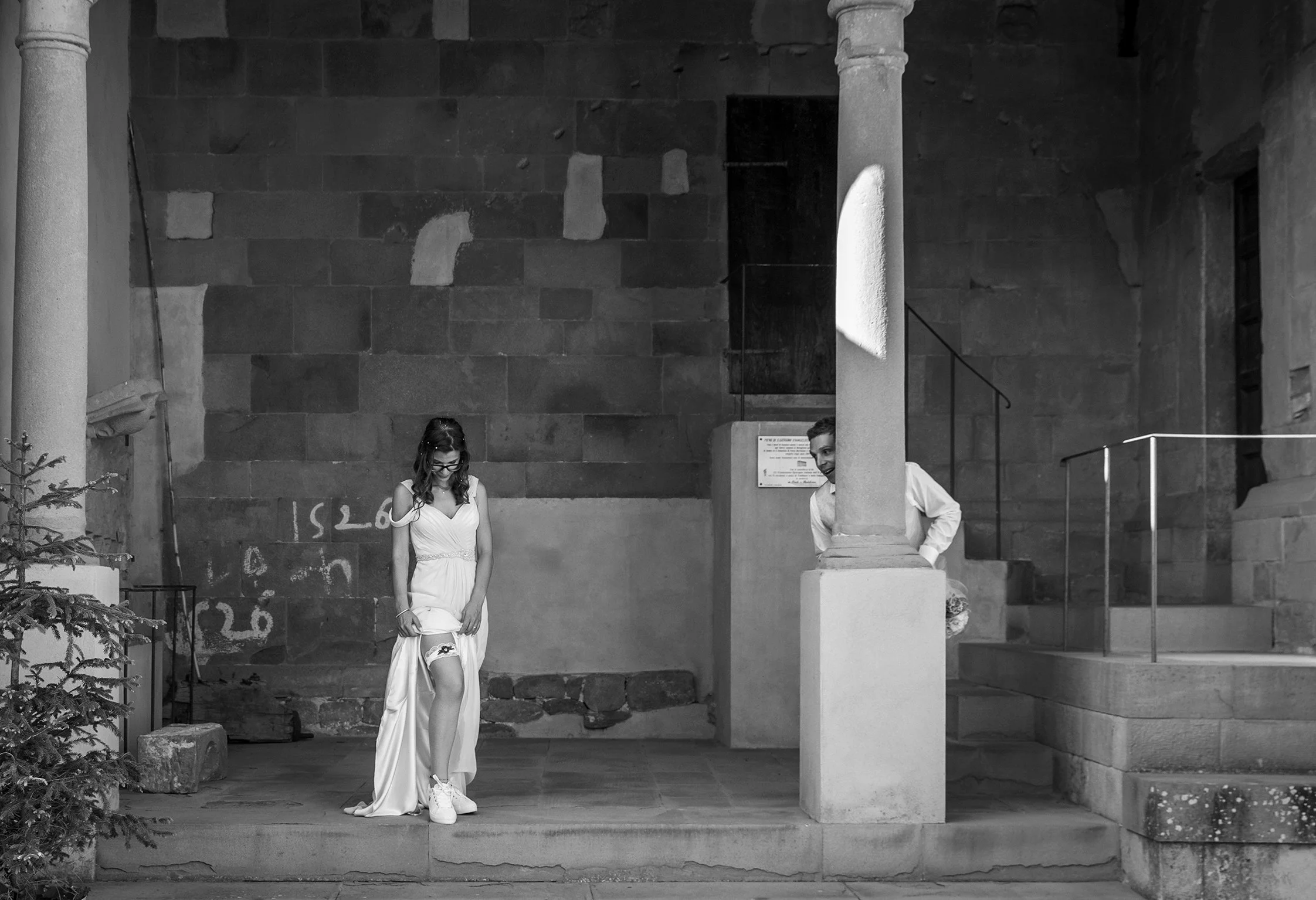 Una donna in abito da sposa mostra la giarrettiera su una gamba, mentre un uomo sbuca da dietro una colonna osservandola. L'immagine è in bianco e nero e si trova in un ambiente architettonico con pareti di pietra.