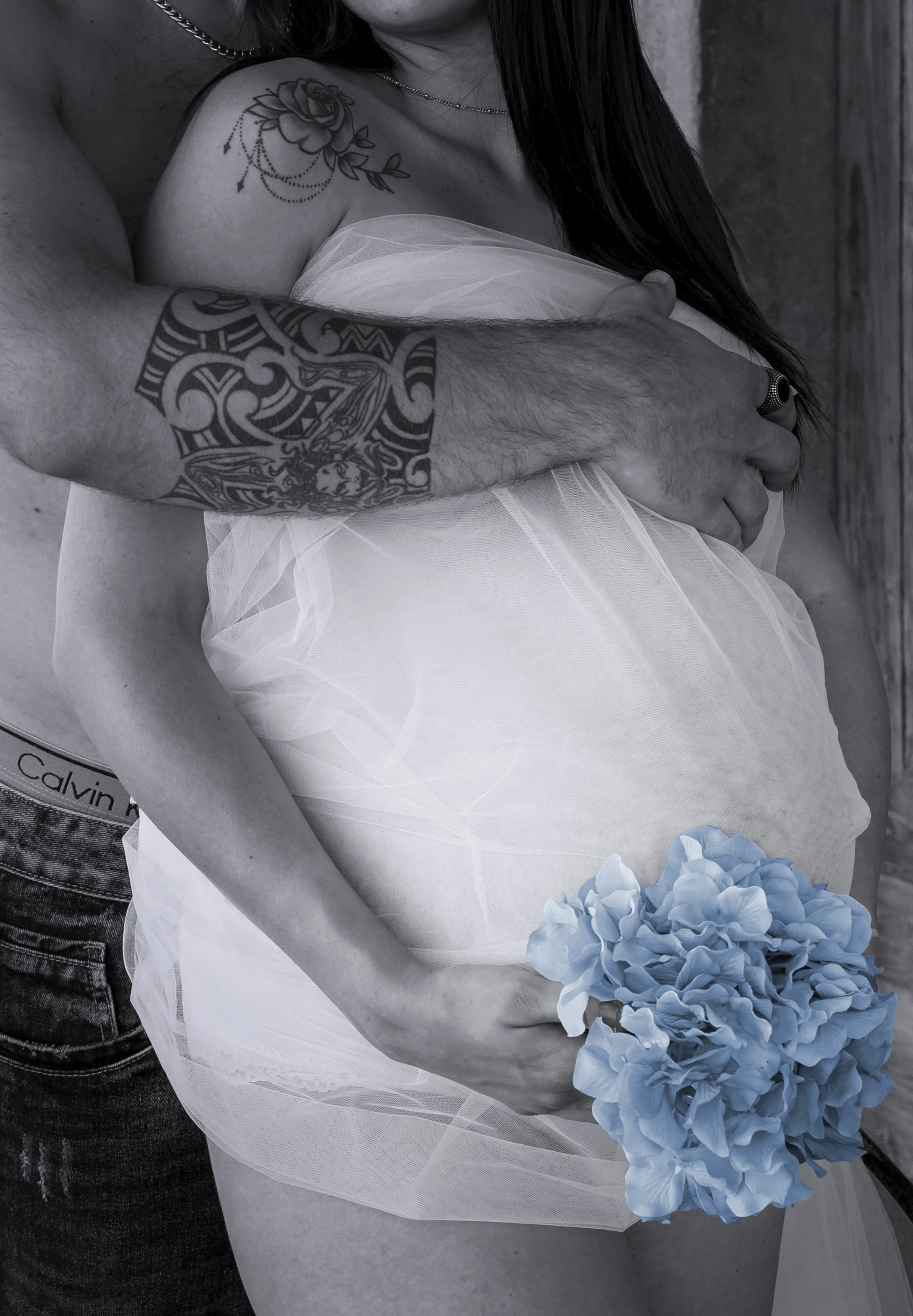 Donna incinta avvolta in tessuto bianco, abbracciata da un uomo con tatuaggio e mazzo di fiori blu.