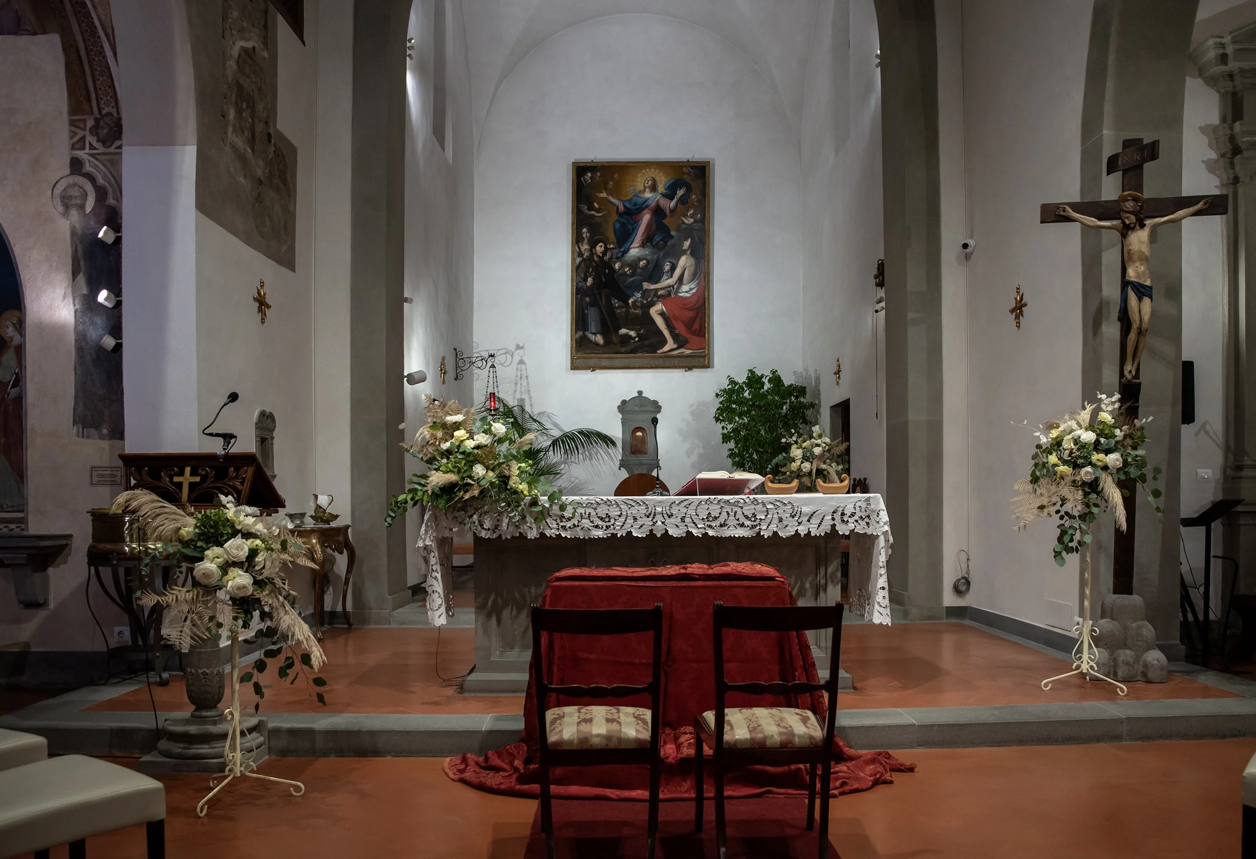 Interno di una chiesa con un altare decorato con fiori, una croce e un dipinto religioso. Due sedie sono posizionate davanti all'altare.