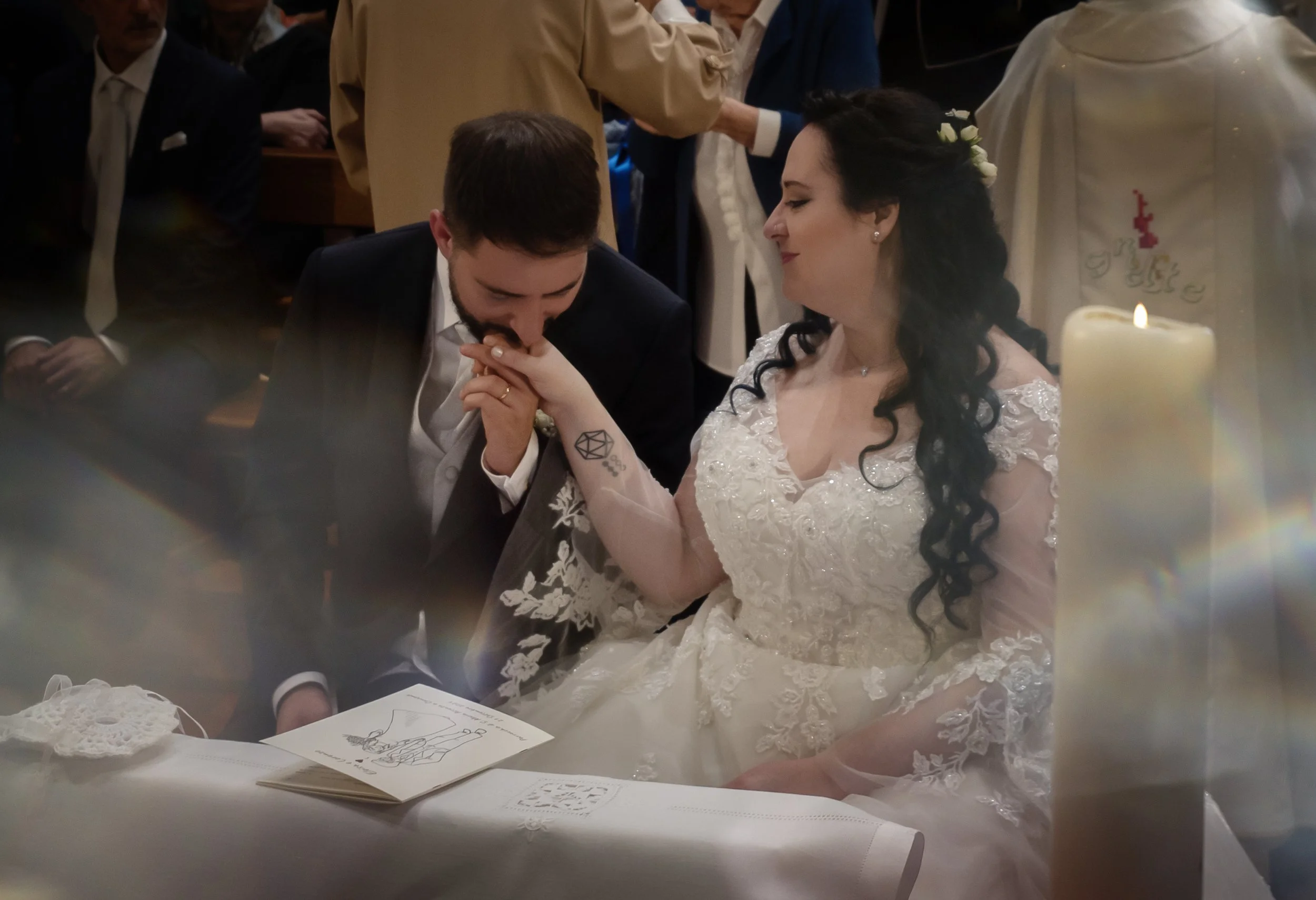 Un matrimonio in chiesa, con gli sposi seduti mentre si tengono per mano. Lo sposo bacia la mano della sposa, che indossa un abito bianco. Una candela accesa è visibile in primo piano.