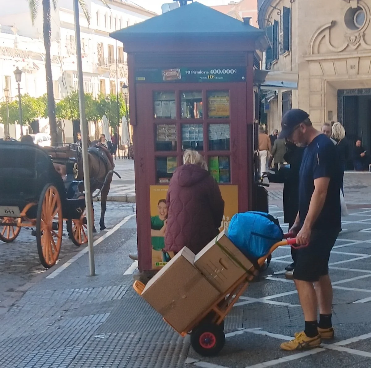 Man pushing a trolley with two boxes and a duffel bag though a busy street, with a horse-drawn carriage in the background