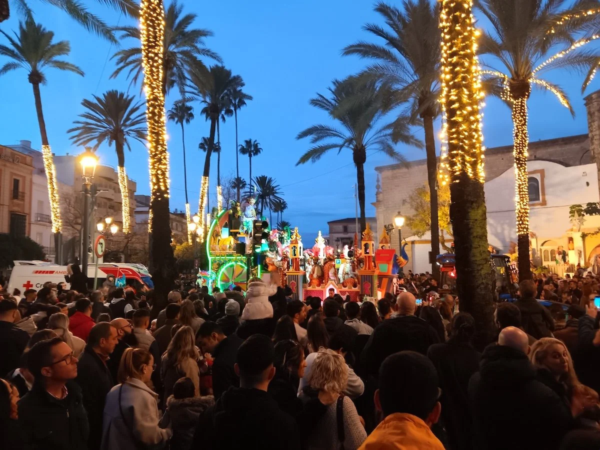 A colourful float among a crowd of people, driving through palm trees and past an old church