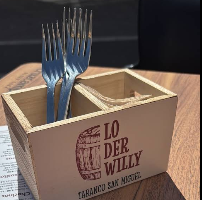 A small wooden box containing two forks and some napkins on an outdoor table. Writing on the box says Lo der Willy..