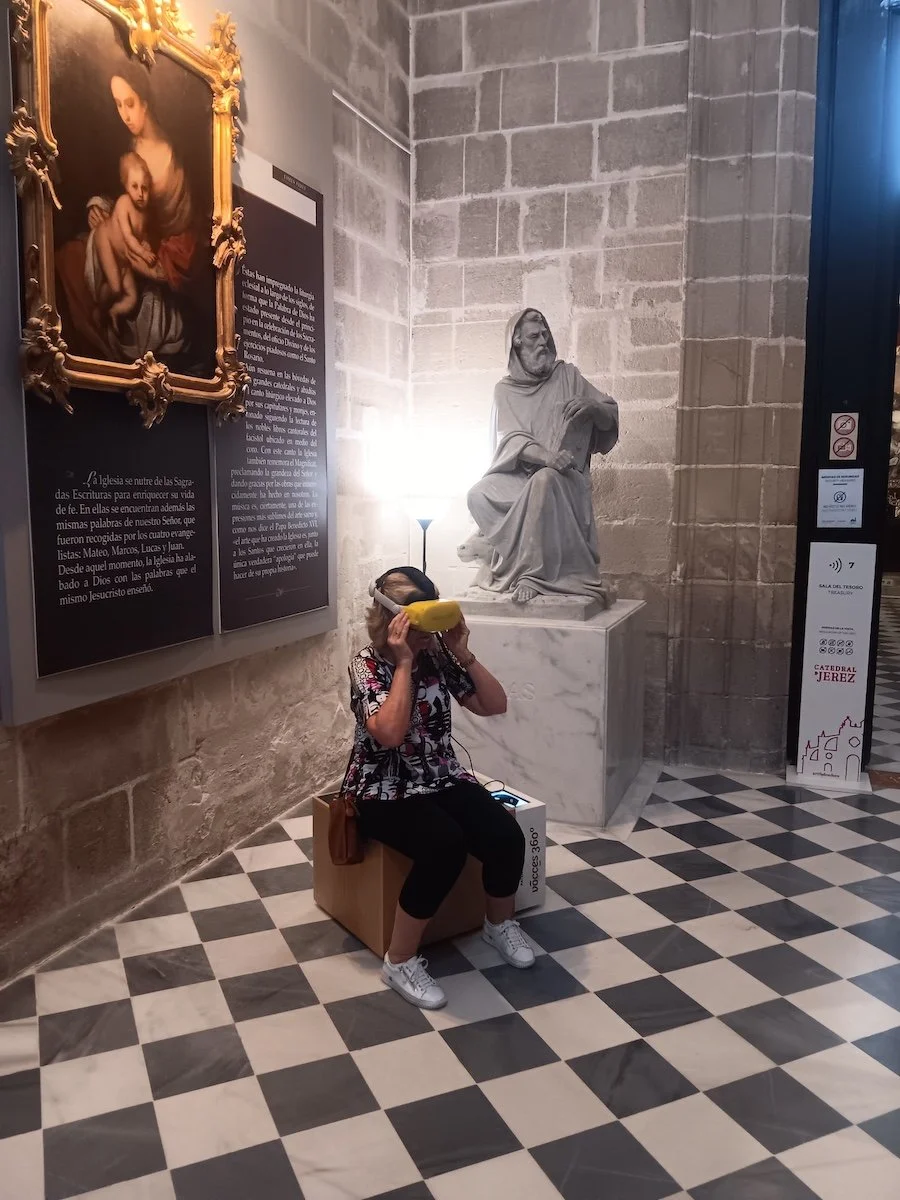 A woman sitting on a stool wearing a virtual reality headset inside a room in a cathedral  with a statue and an old painting
